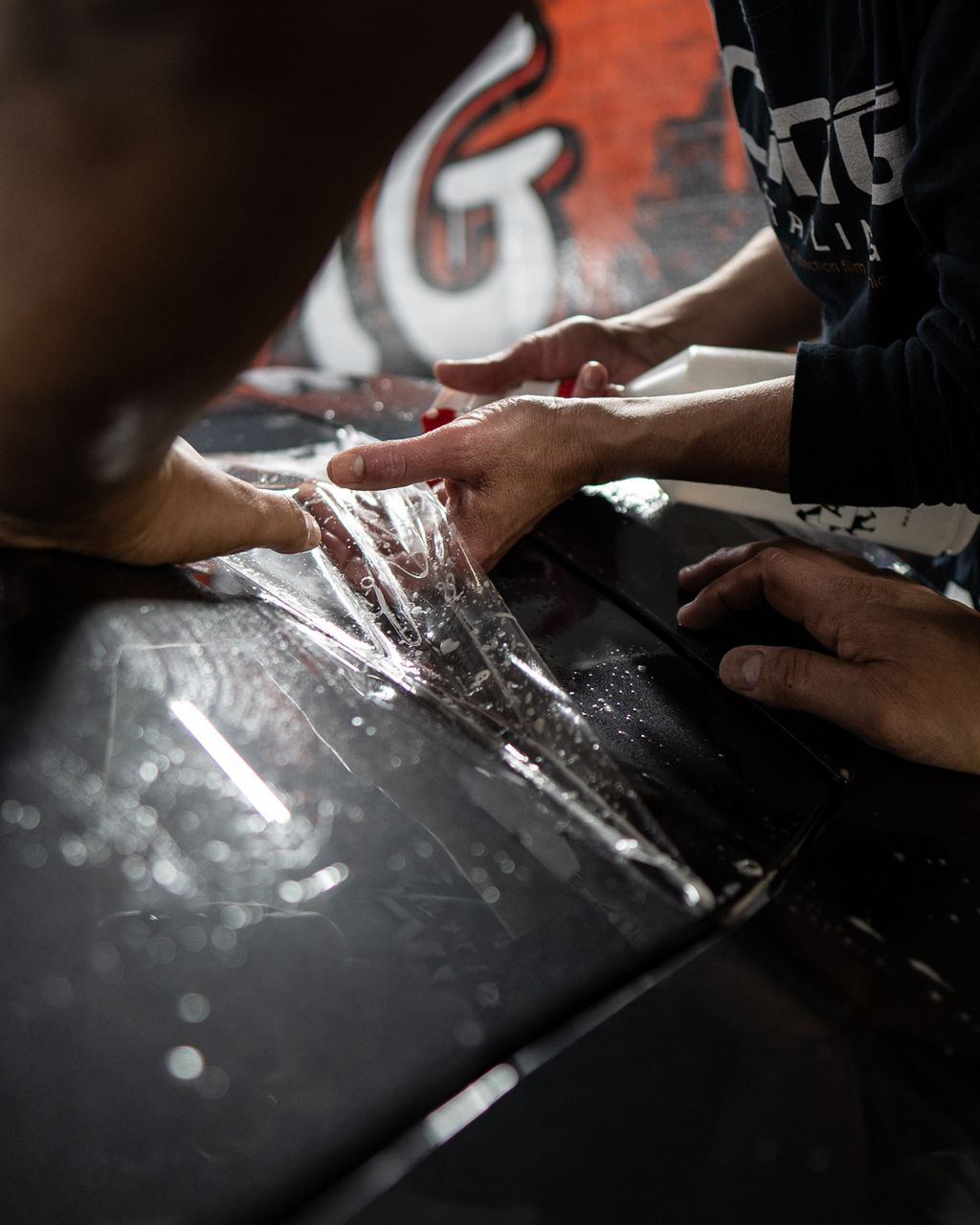 Hands applying a clear film to a dark surface, possibly a car.