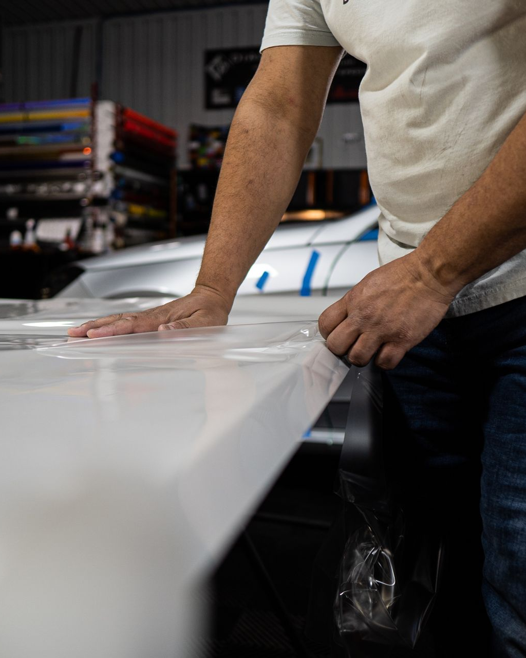 Person applying film to a white surface, possibly a car. Shop interior with rolls of colored material.