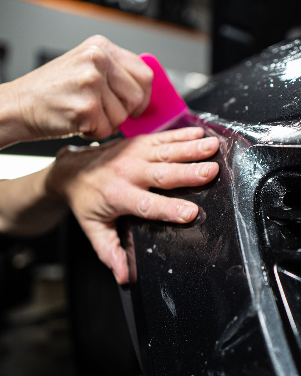 Person using a pink squeegee to apply a film to a black car bumper.
