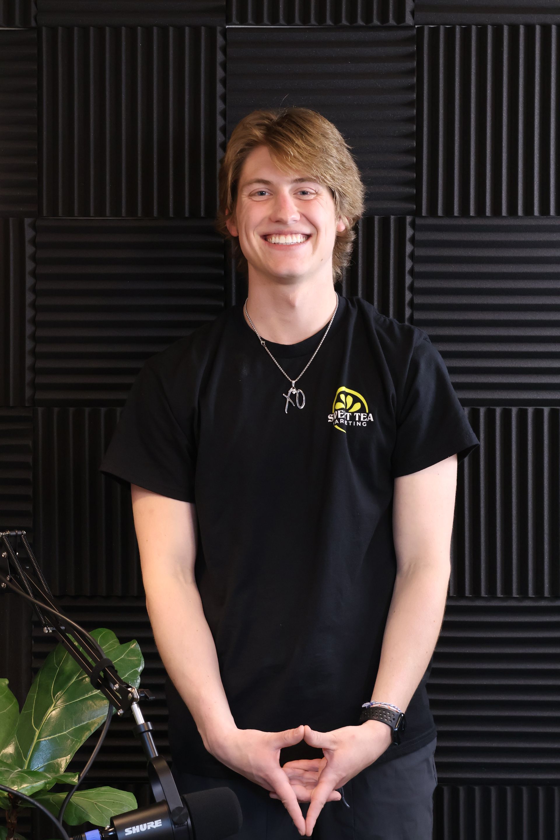 A young man wearing a black t-shirt and a necklace is standing in front of a microphone.