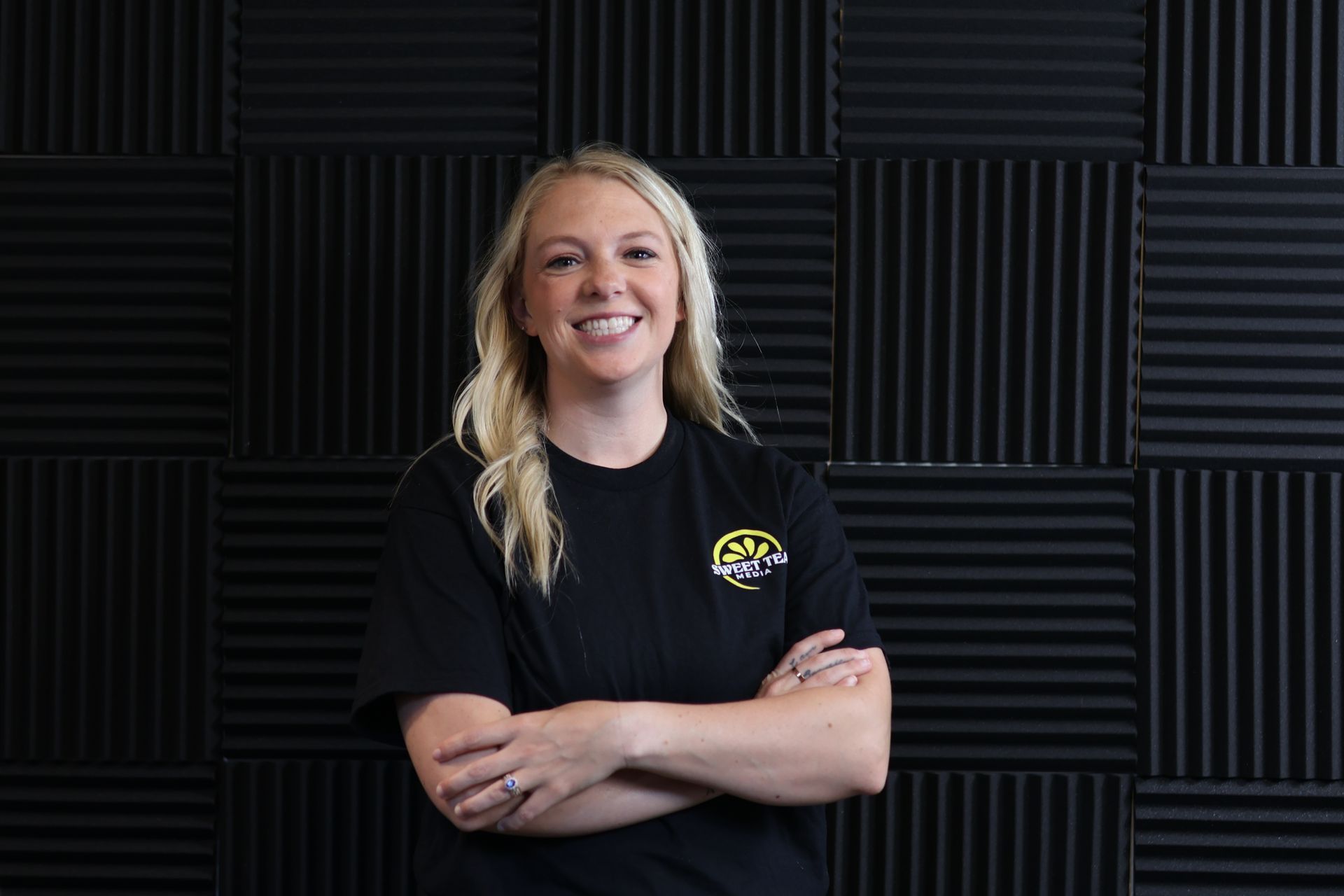 A woman in a black shirt is standing in front of a black wall with her arms crossed.