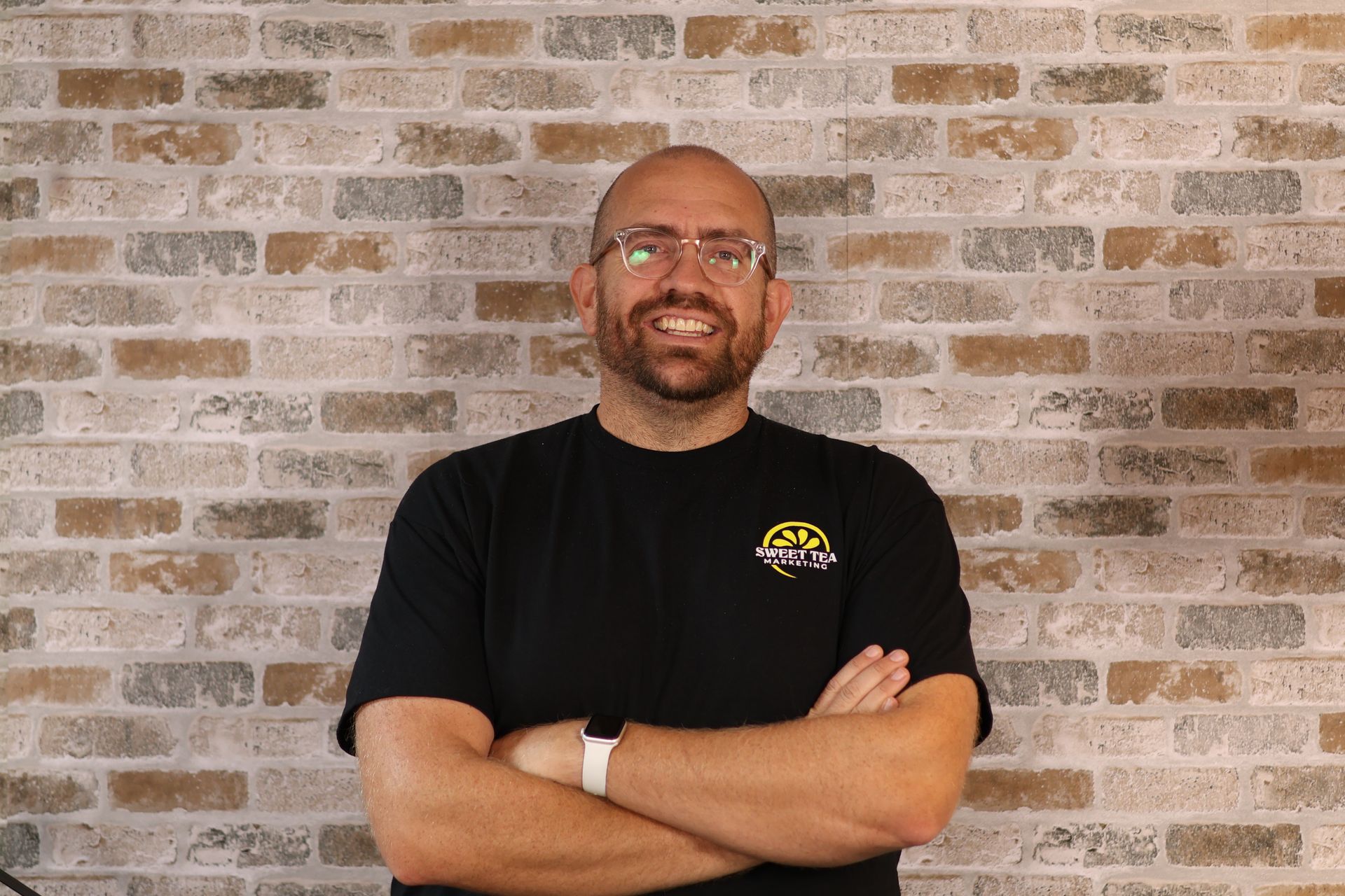 A bald man with glasses is standing in front of a brick wall with his arms crossed.