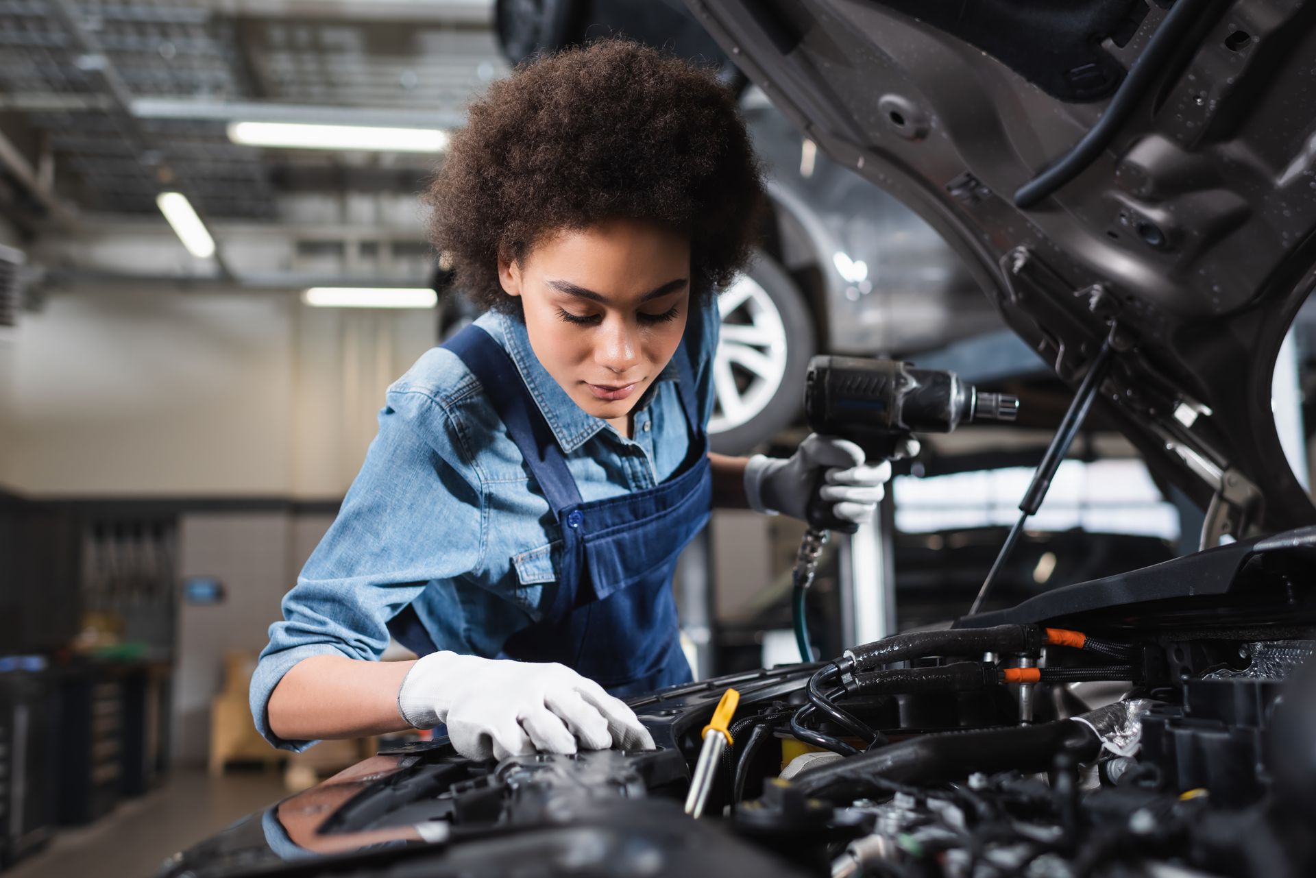 A car mechanic in overalls holding an electric screwdriver and working with a car motor at a garage.