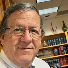 A man wearing glasses and a tie is standing in front of a bookshelf.