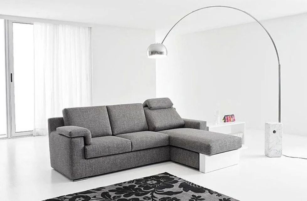 Divano componibile grigio con chaise longue annessa in una stanza bianca; lampada da terra ad arco.