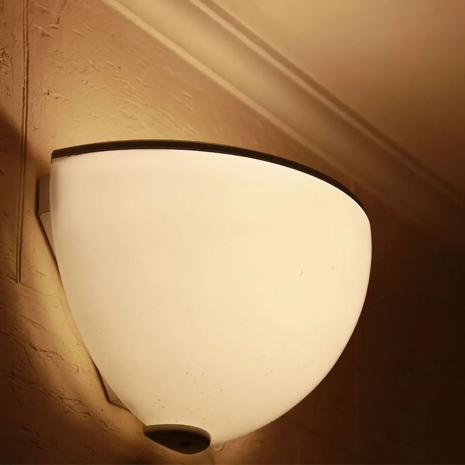 Lampada da parete a forma di lacrima con paralume bianco opaco e finiture nere, che illumina una parete beige.