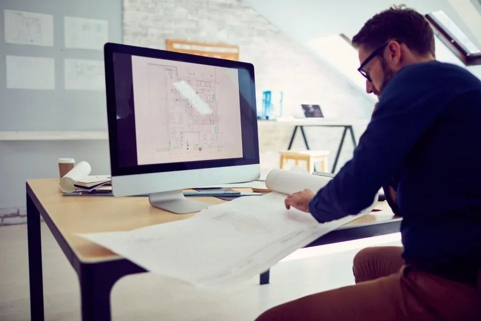 Uomo che esamina progetti architettonici alla scrivania con monitor di computer, ambiente interno.