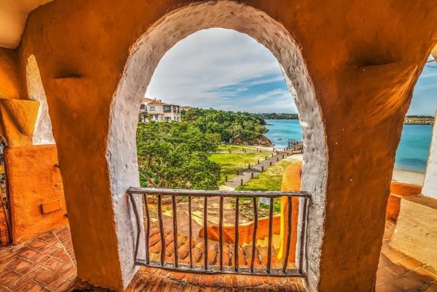 Vista attraverso una finestra ad arco: paesaggio costiero con alberi, acqua ed edifici