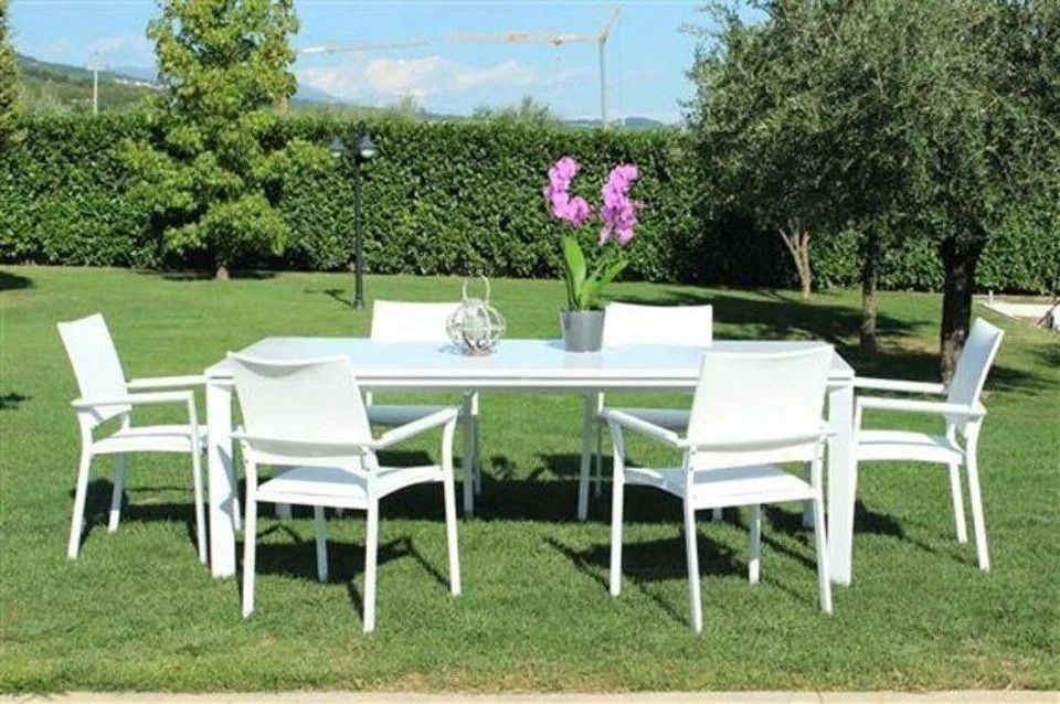 Tavolo da pranzo bianco all'aperto su prato verde con una siepe sullo sfondo.