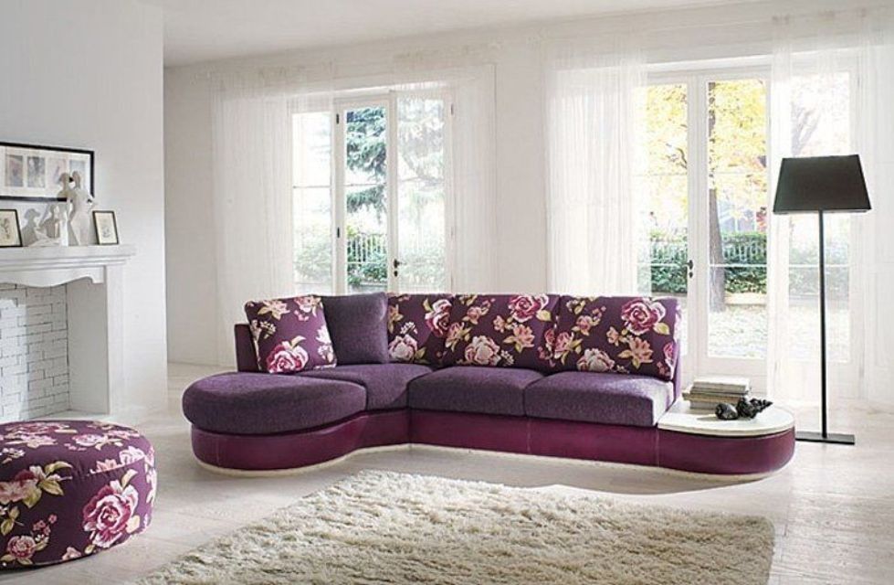 Divano componibile con motivi floreali viola in un luminoso soggiorno, pouf, finestre e una lampada da terra.
