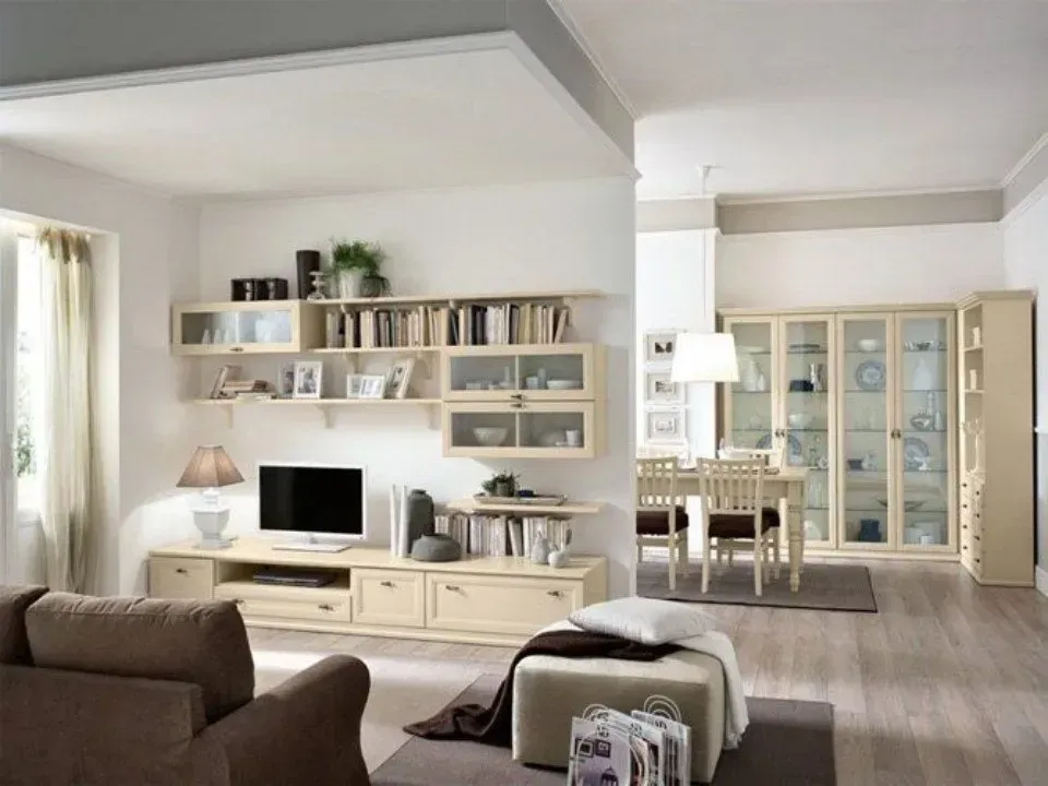 Soggiorno con mobili beige, TV e scaffali; zona pranzo visibile attraverso un'apertura.