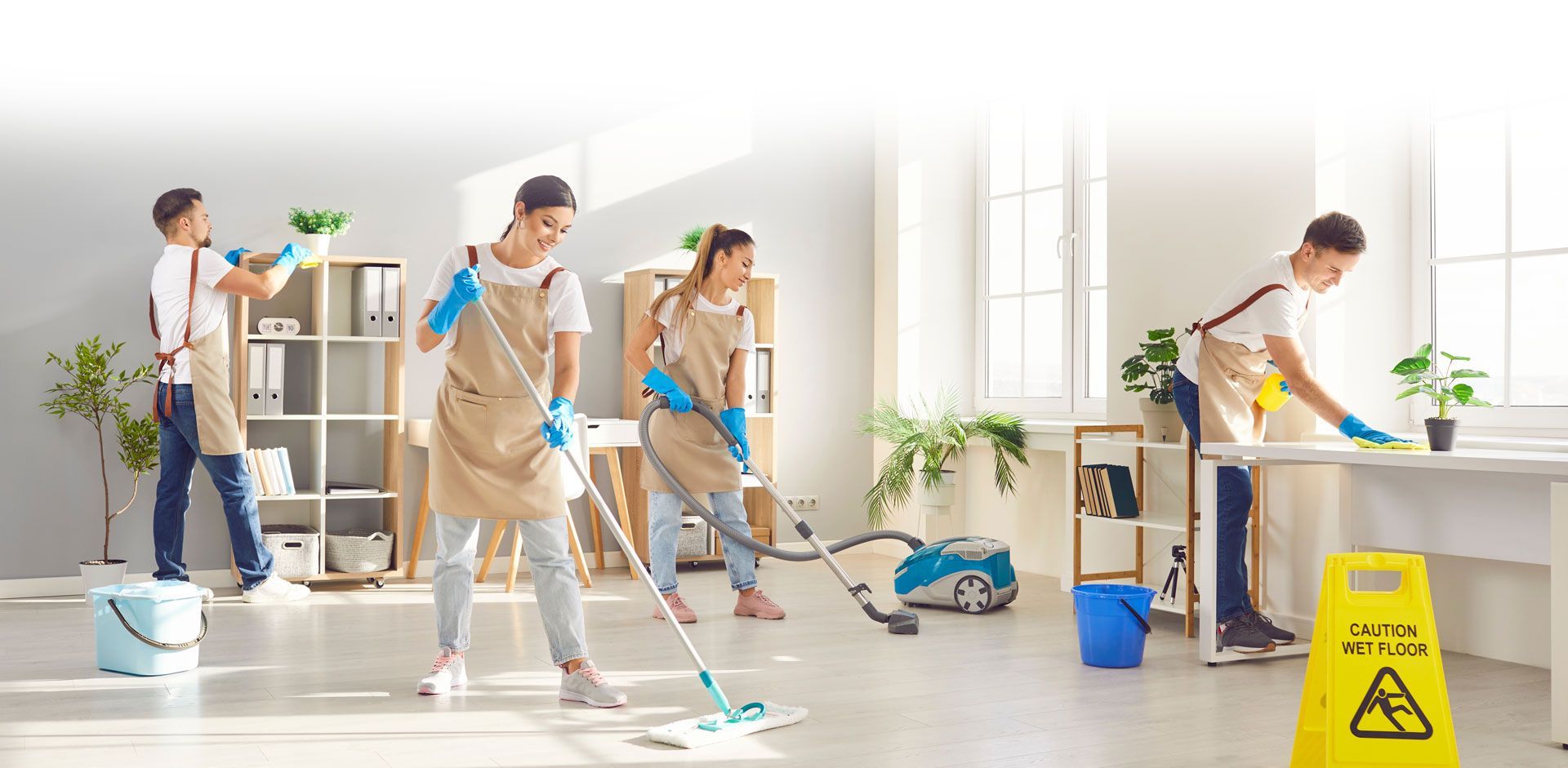 A group of cleaners cleaning an office. One mops, one vacuums, one dusts, and one puts plants away.