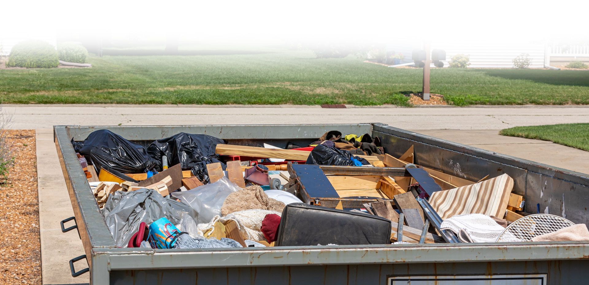 A large dumpster overflowing with assorted trash, sitting outdoors near grass and a sidewalk.