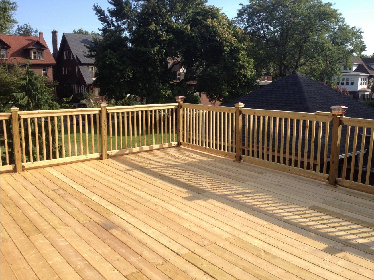 Wooden deck with railing, bathed in sunlight, overlooking neighborhood with houses and trees.