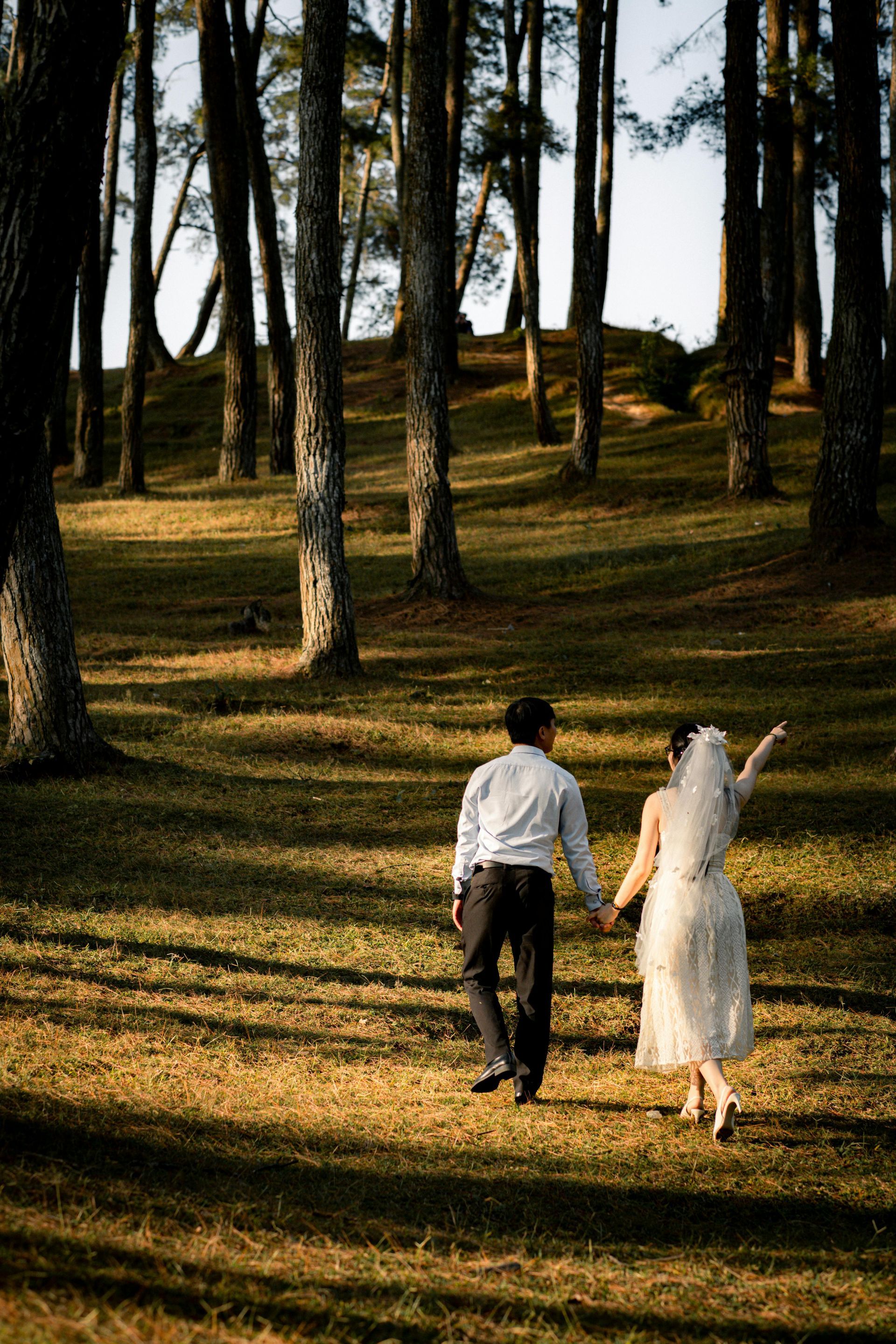 A bride and groom are walking through a forest holding hands.
