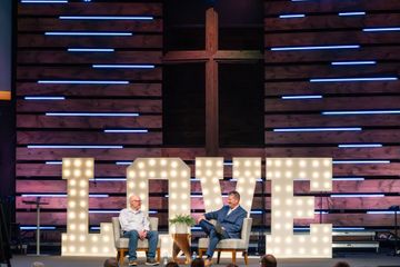 Two men are sitting in front of a large sign that says love.