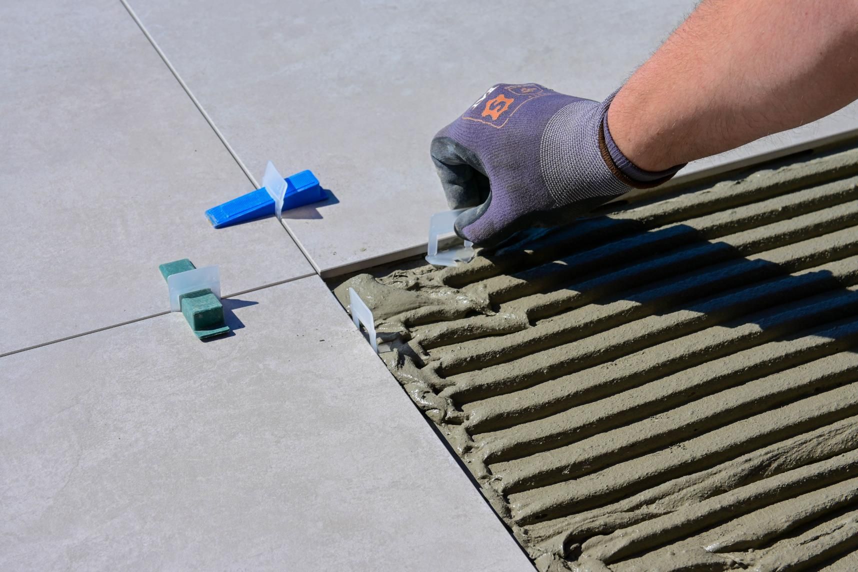 Gloved hand spreading mortar on floor tiles beside blue and green spacers