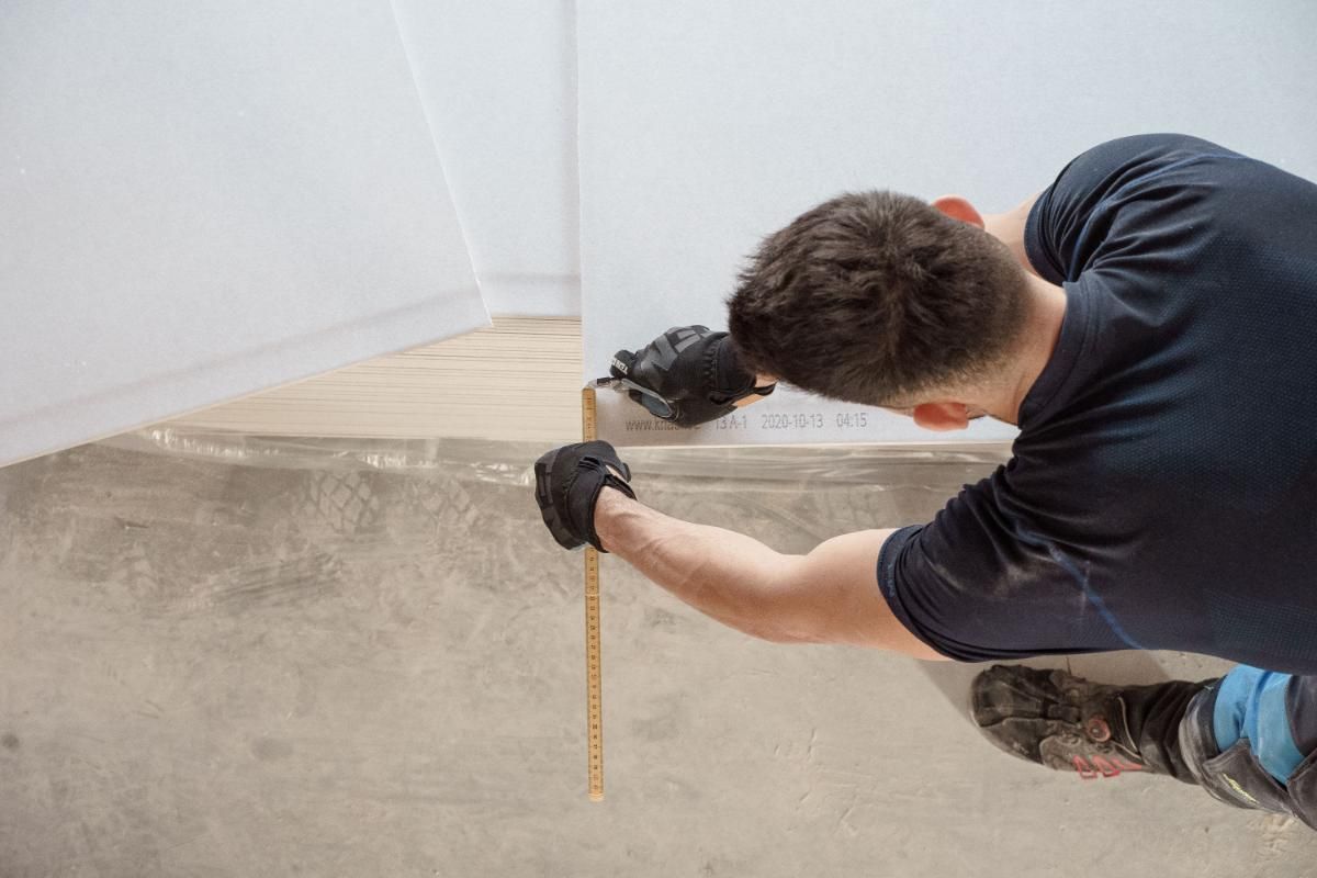 Worker measuring concrete floor edge with a tape measure while kneeling indoors.