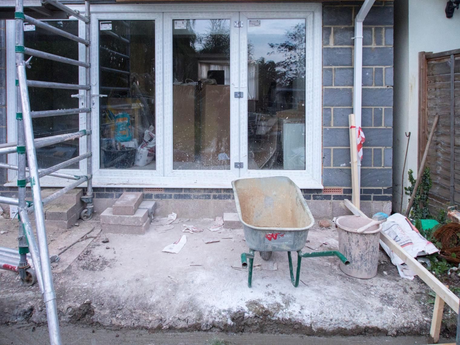 Partially built patio with a wheelbarrow in front of sliding glass doors, surrounded by construction debris