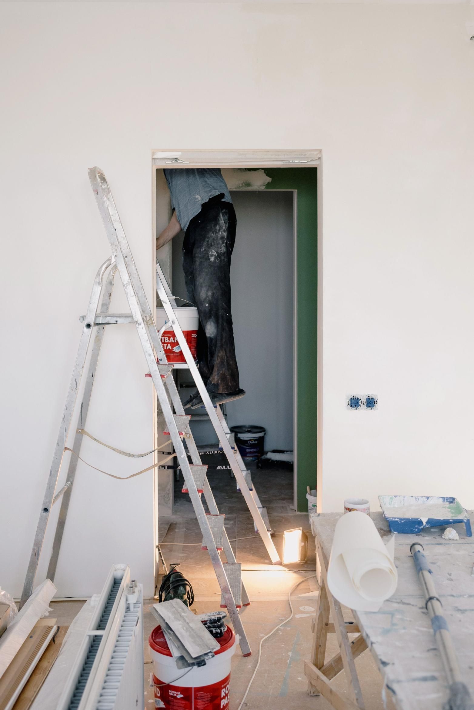 Ladders and painting supplies in a white room with an open doorway under renovation.