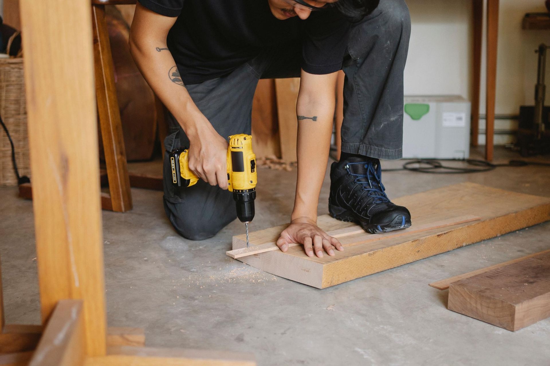 Person kneels drilling a wood board on a floor, with tools and lumber nearby.