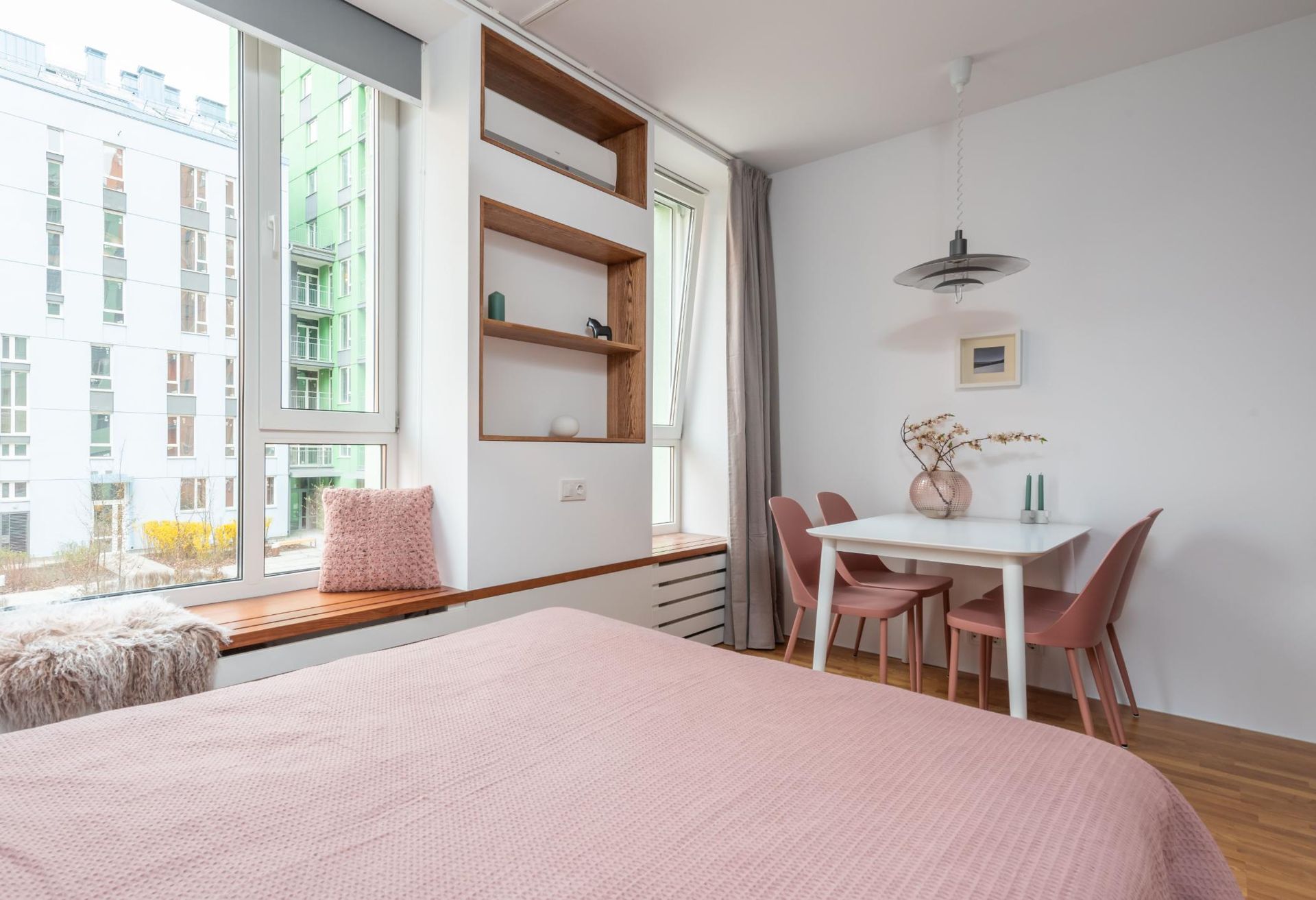 Bright bedroom with pink bedspread, large windows, built-in shelves, and a small dining table by the wall