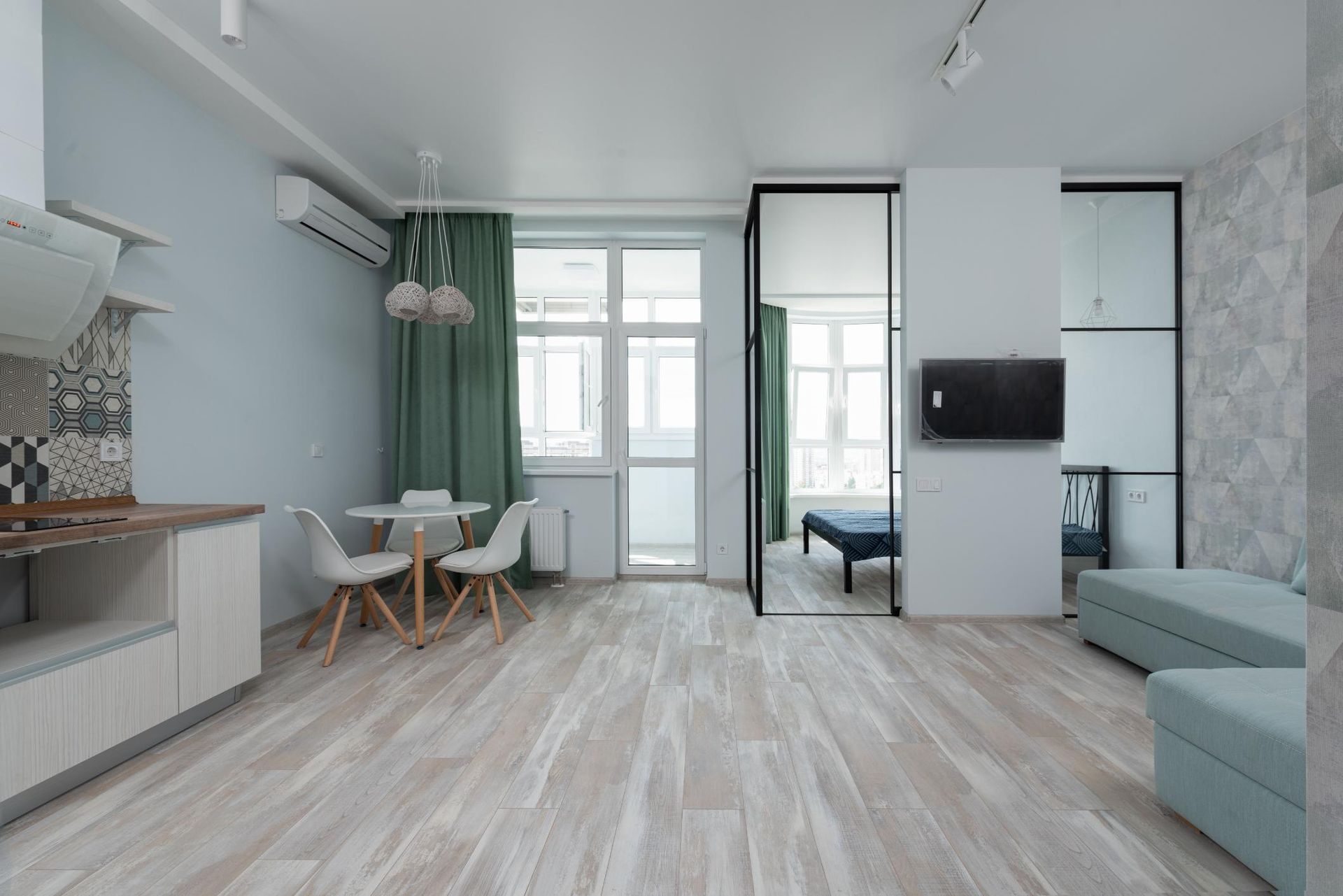 Modern studio apartment with light wood floors, green curtains, small dining table, sofa, and wall-mounted TV