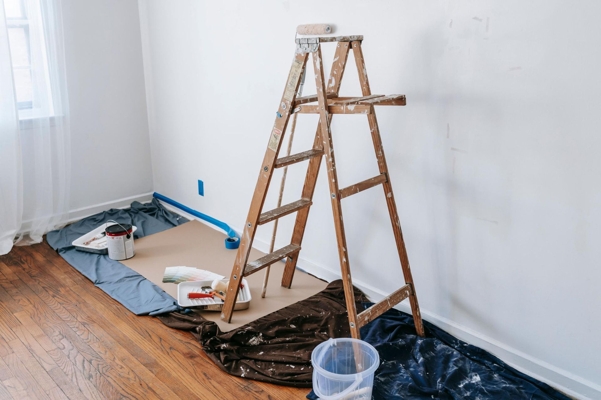 Paint supplies and a wooden step ladder in a room with drop cloths and partially painted walls