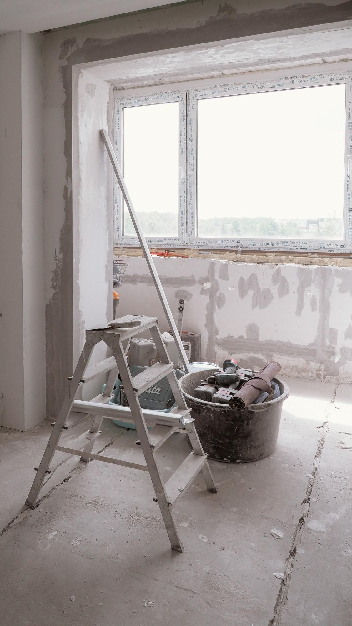 Renovation room with ladder, bucket, and unfinished plaster walls by a window