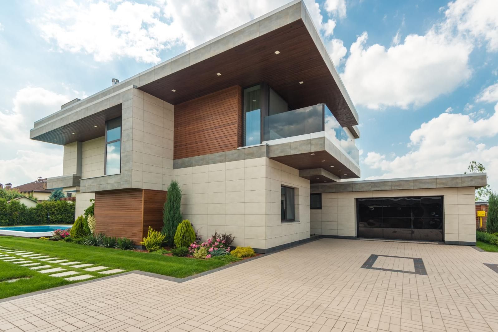 Modern two-story house with wood accents, balcony, and a large driveway under a cloudy sky