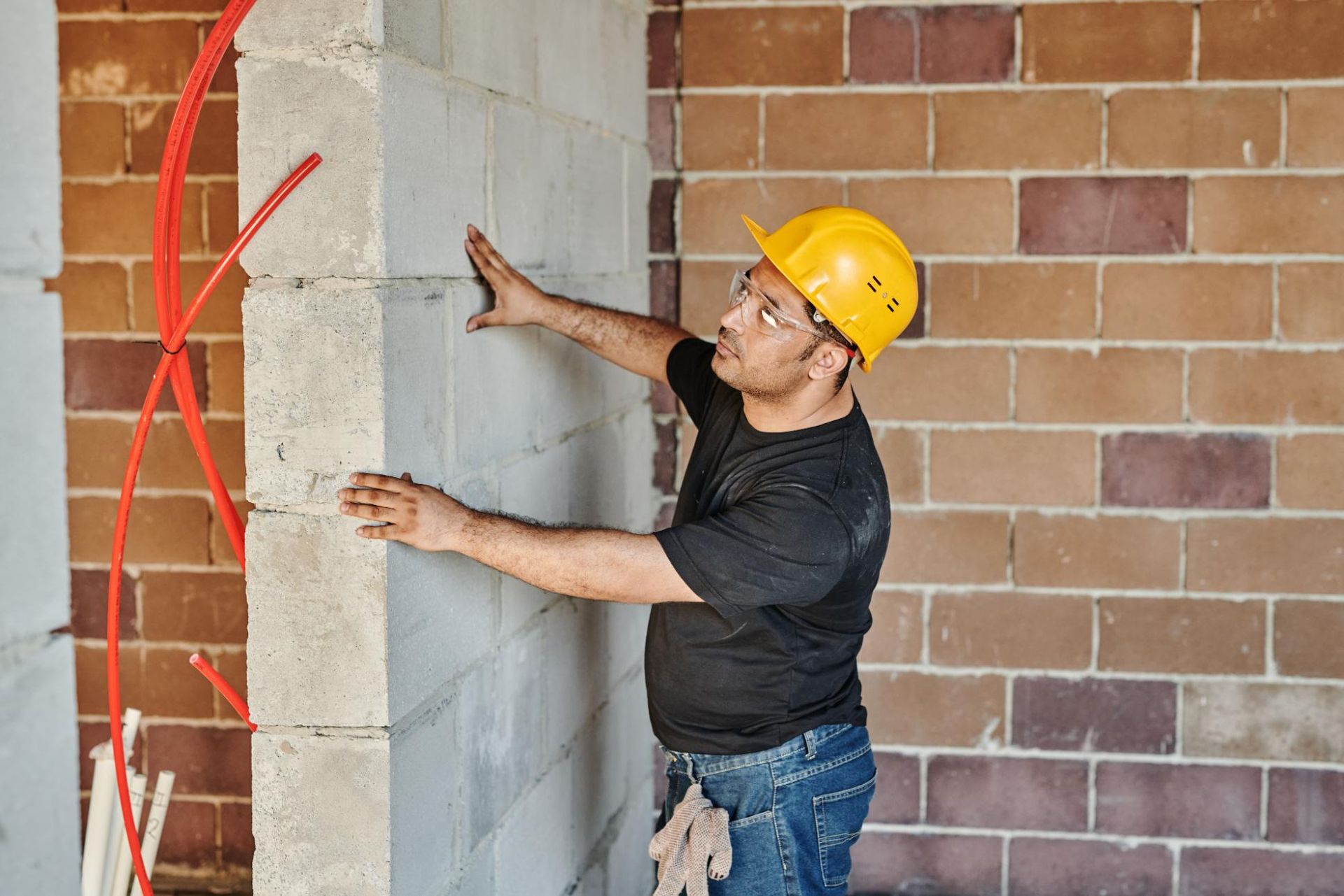 Worker in a yellow hard hat measuring a cinder block wall inside a brick building.