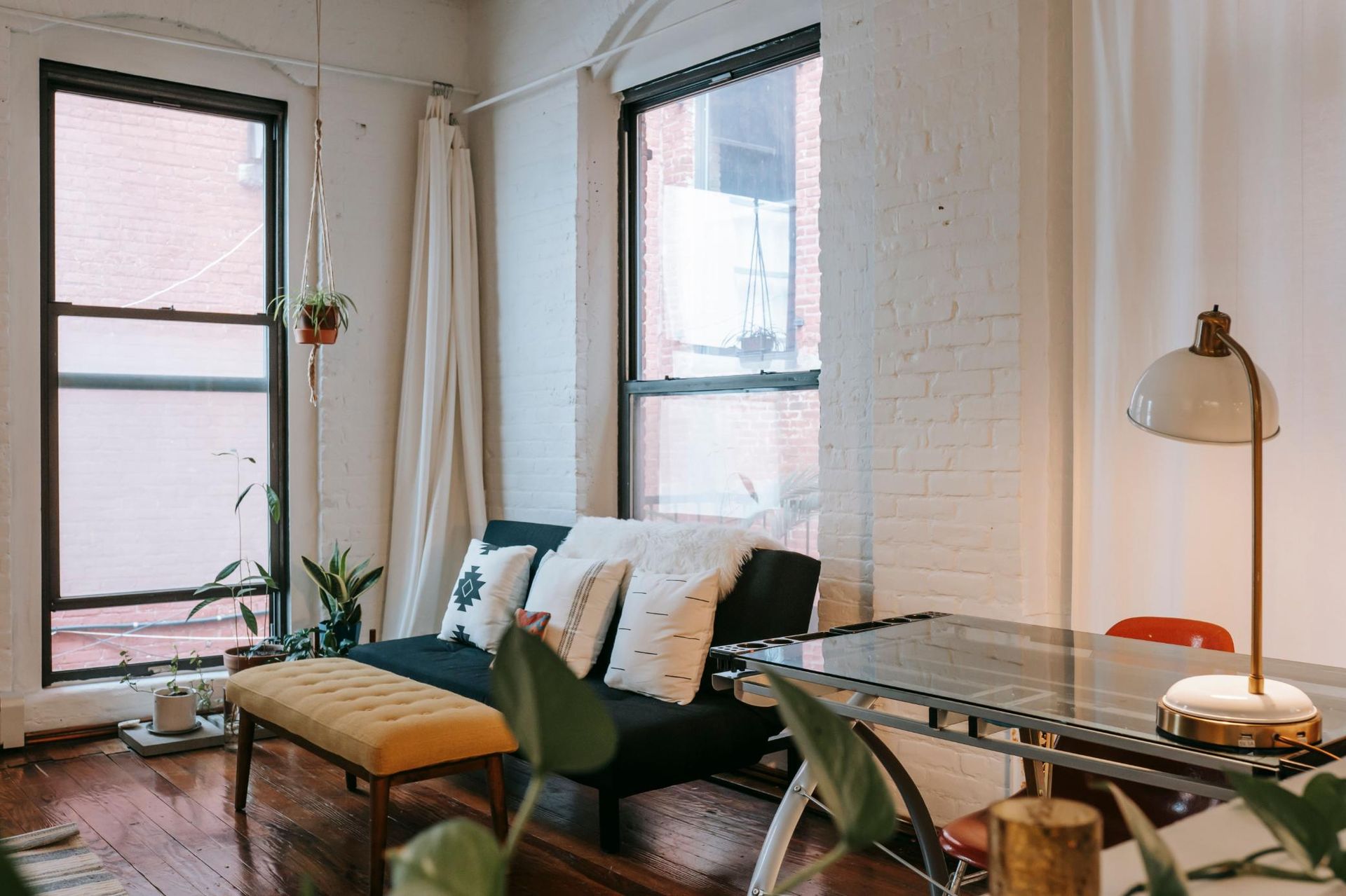 Bright living room with black sofa, tan bench, desk lamp, and tall windows with blinds