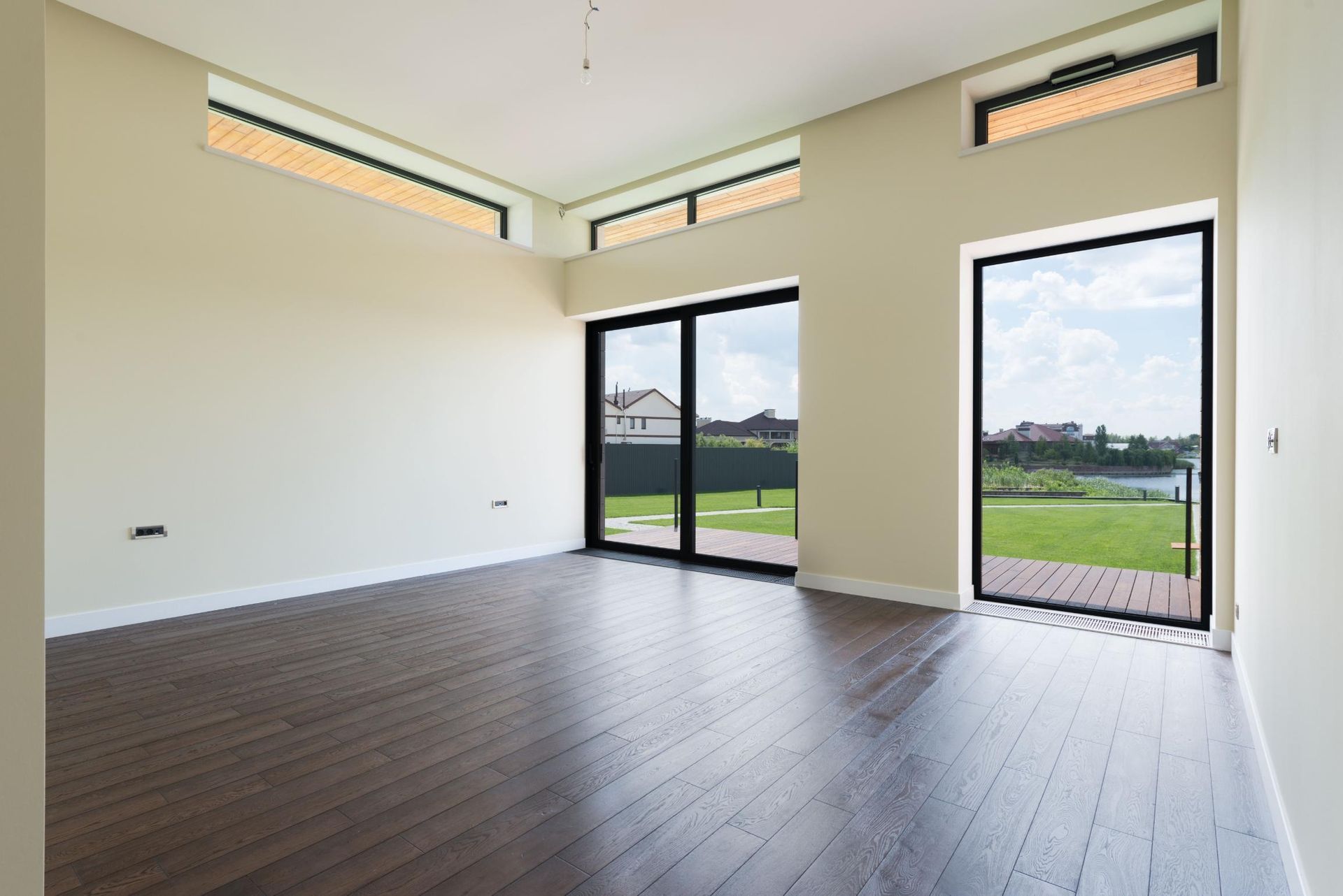 Empty modern room with dark wood floors, white walls, and tall windows and glass doors overlooking greenery