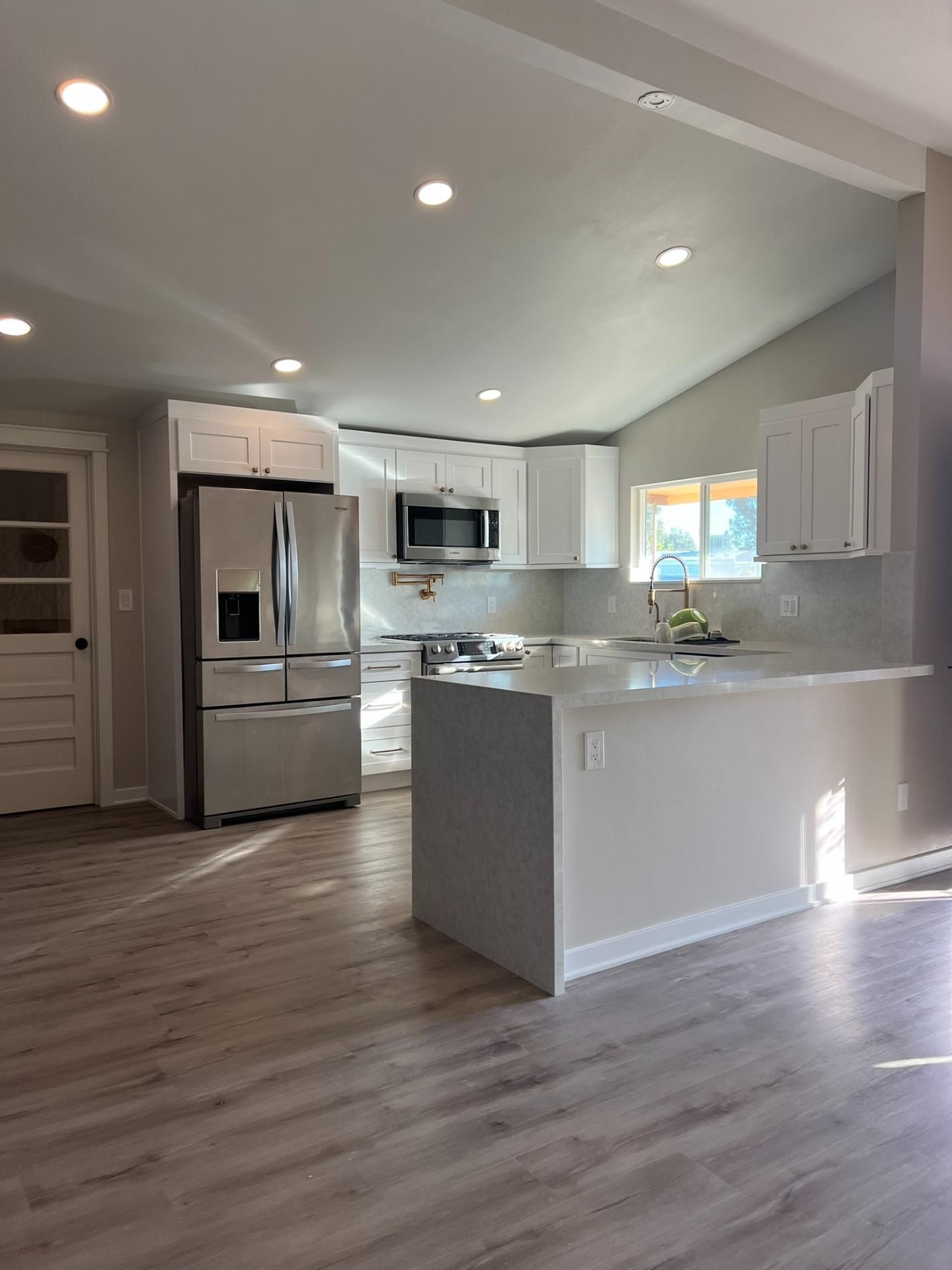 Bright modern kitchen with stainless steel appliances, white cabinets, island, and gray wood-look floors.