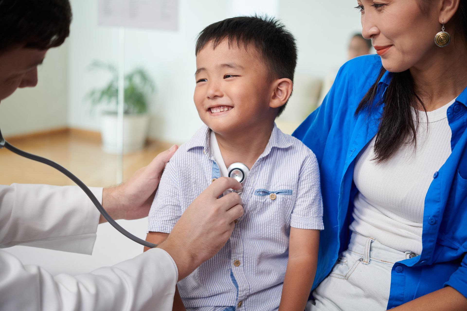 Doctor using a stethoscope on a smiling boy's chest; mother sits beside them.