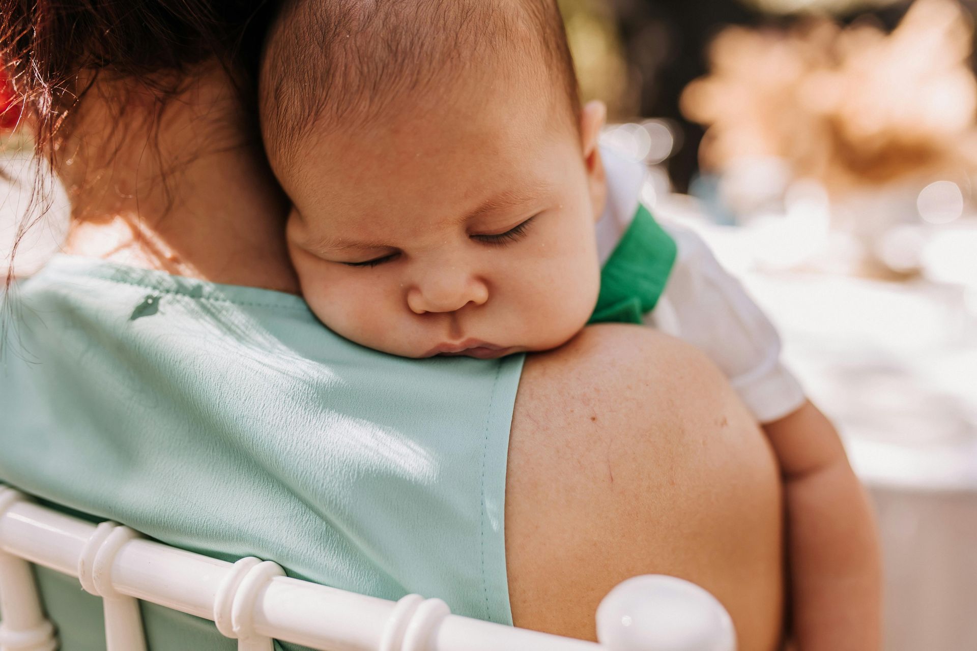 Sleeping baby resting on a person's shoulder, wearing white shirt with green details, outdoors.