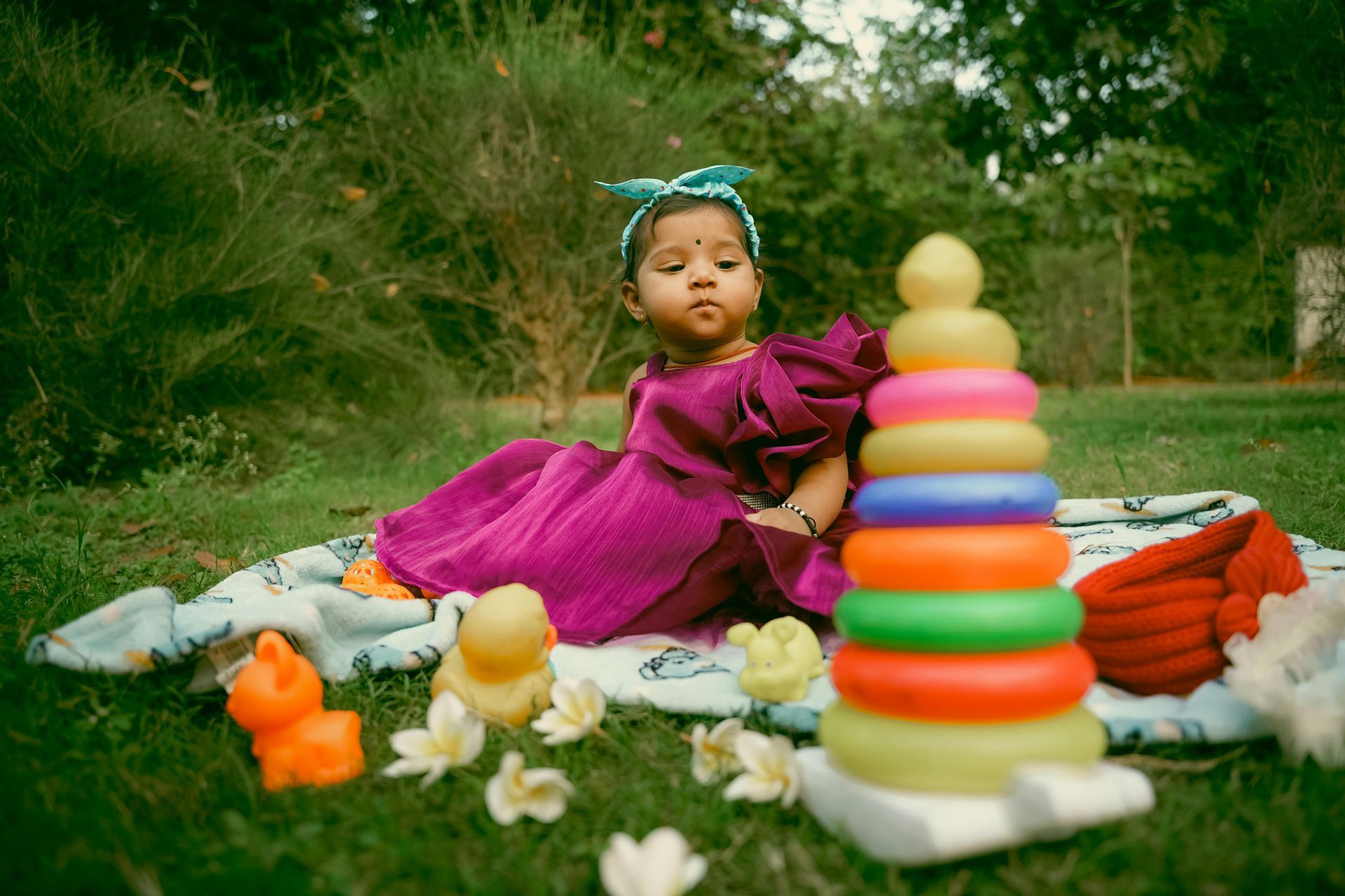 Baby sitting on a blanket outdoors, wearing a purple dress and headband, with colorful toys.