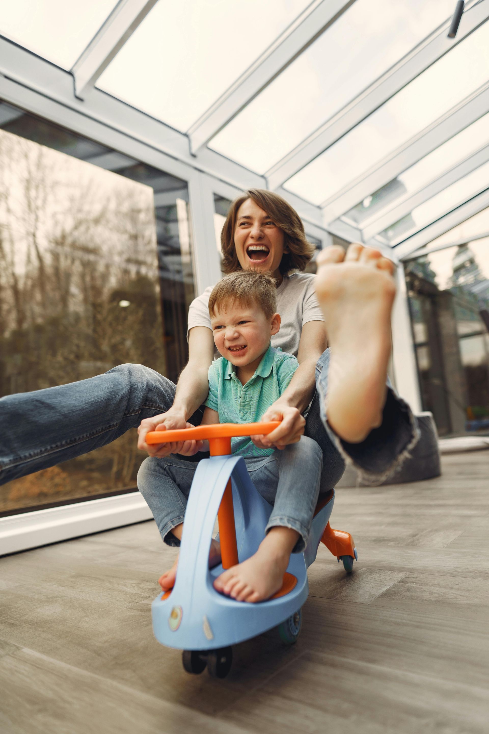 Woman and child laughing, riding a blue and orange toy car indoors; woman's foot up.