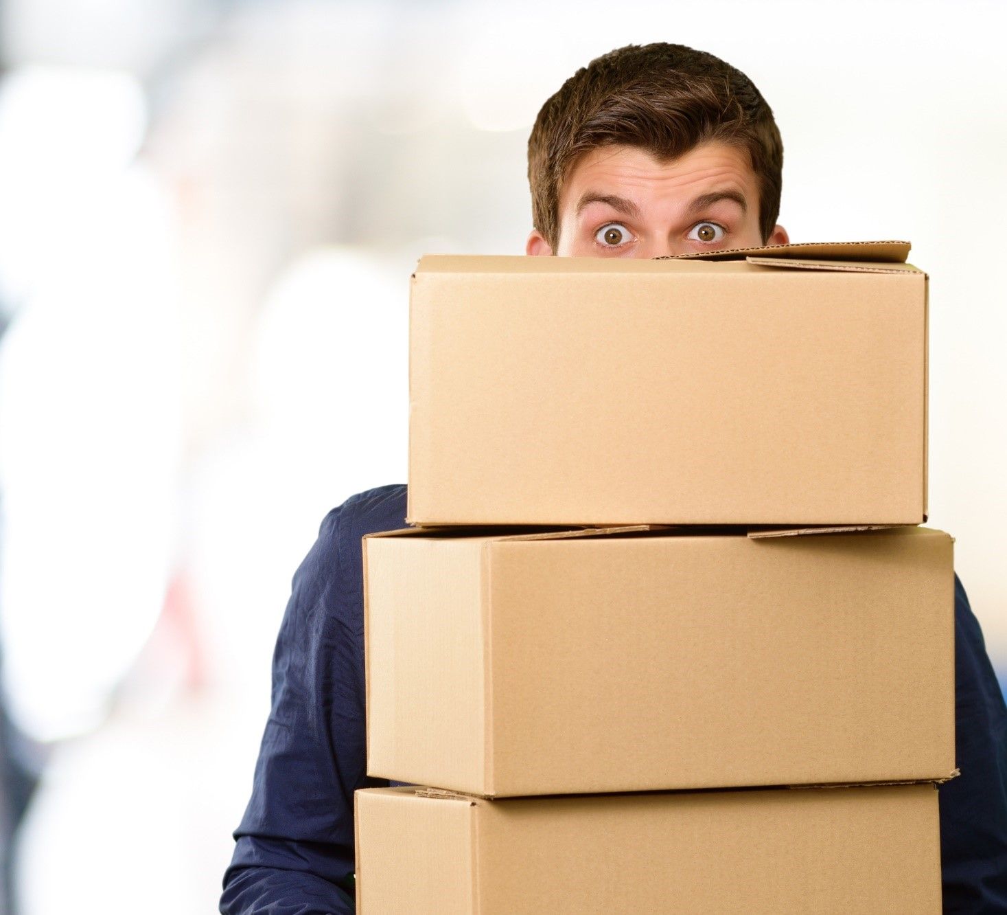 A man wearing a blue jacket with a surprised look on his face, moving 3 brown cardboard boxes.