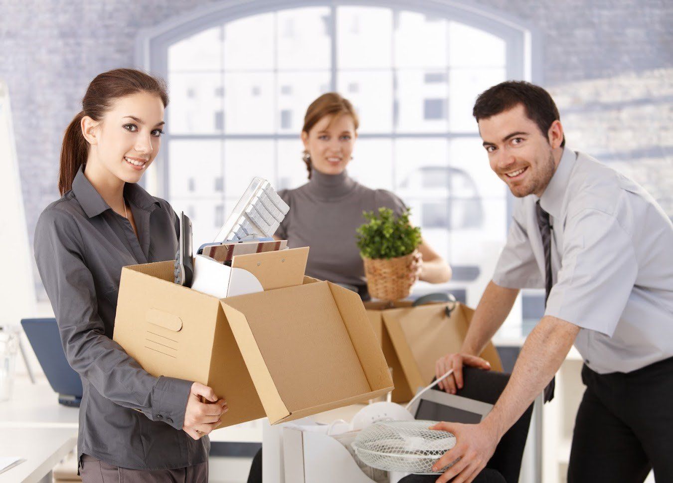 A team of movers, featuring two women holding boxes and a man, packing away office furniture together.