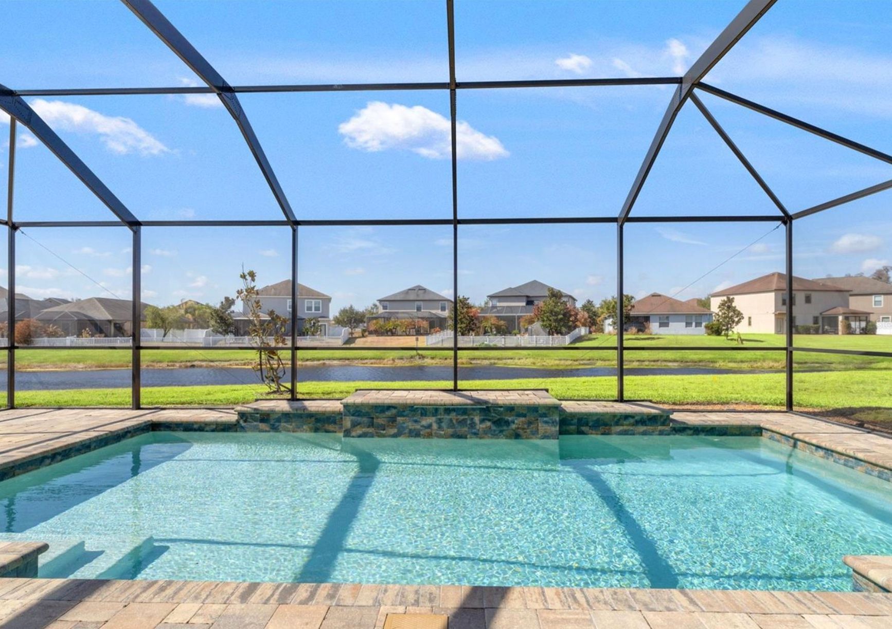 Swimming pool with screened enclosure, blue water, and houses in the background under a blue sky.