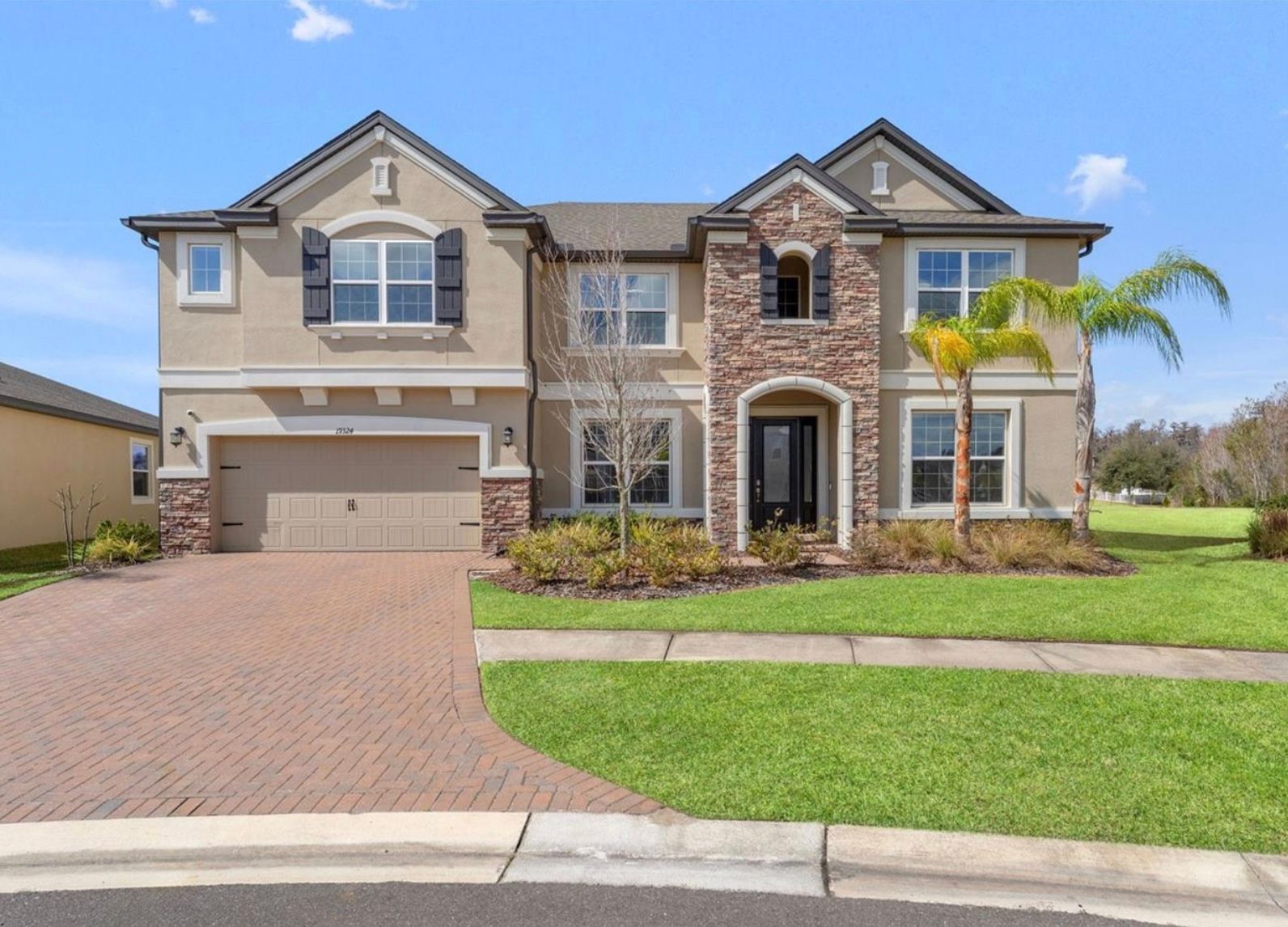 Two-story house with tan stucco, brick accents, and a paved driveway under a blue sky.
