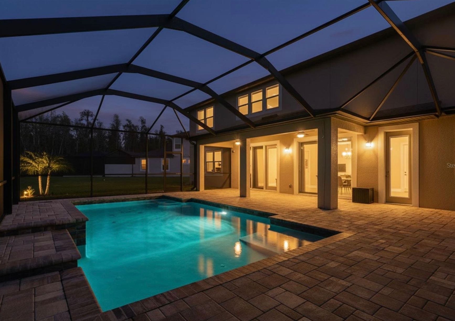 Pool area at night with lit pool, patio, and house in the background under a screened enclosure.