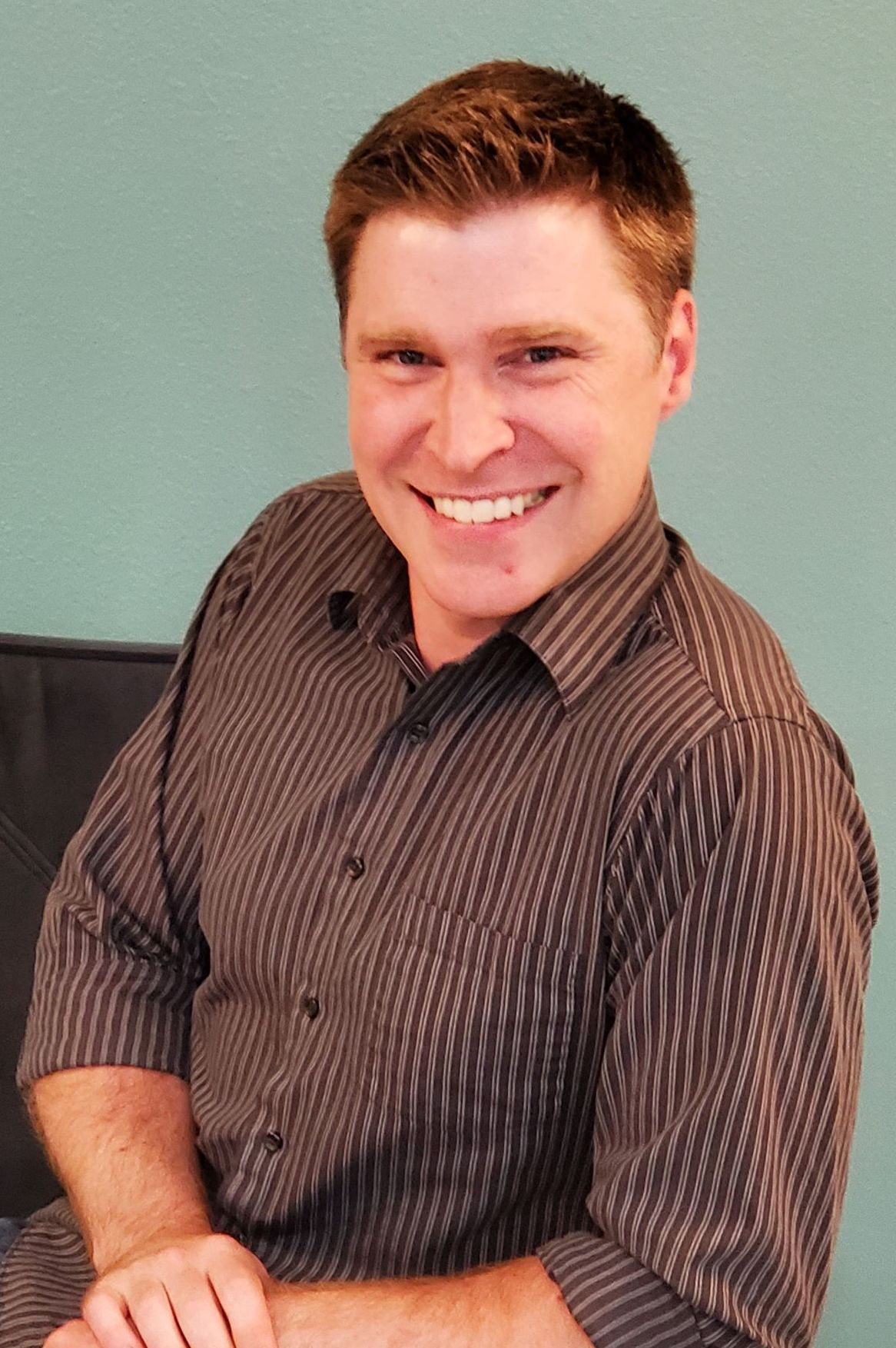 A man in a striped brown dress shirt and short brown hair, smiling.