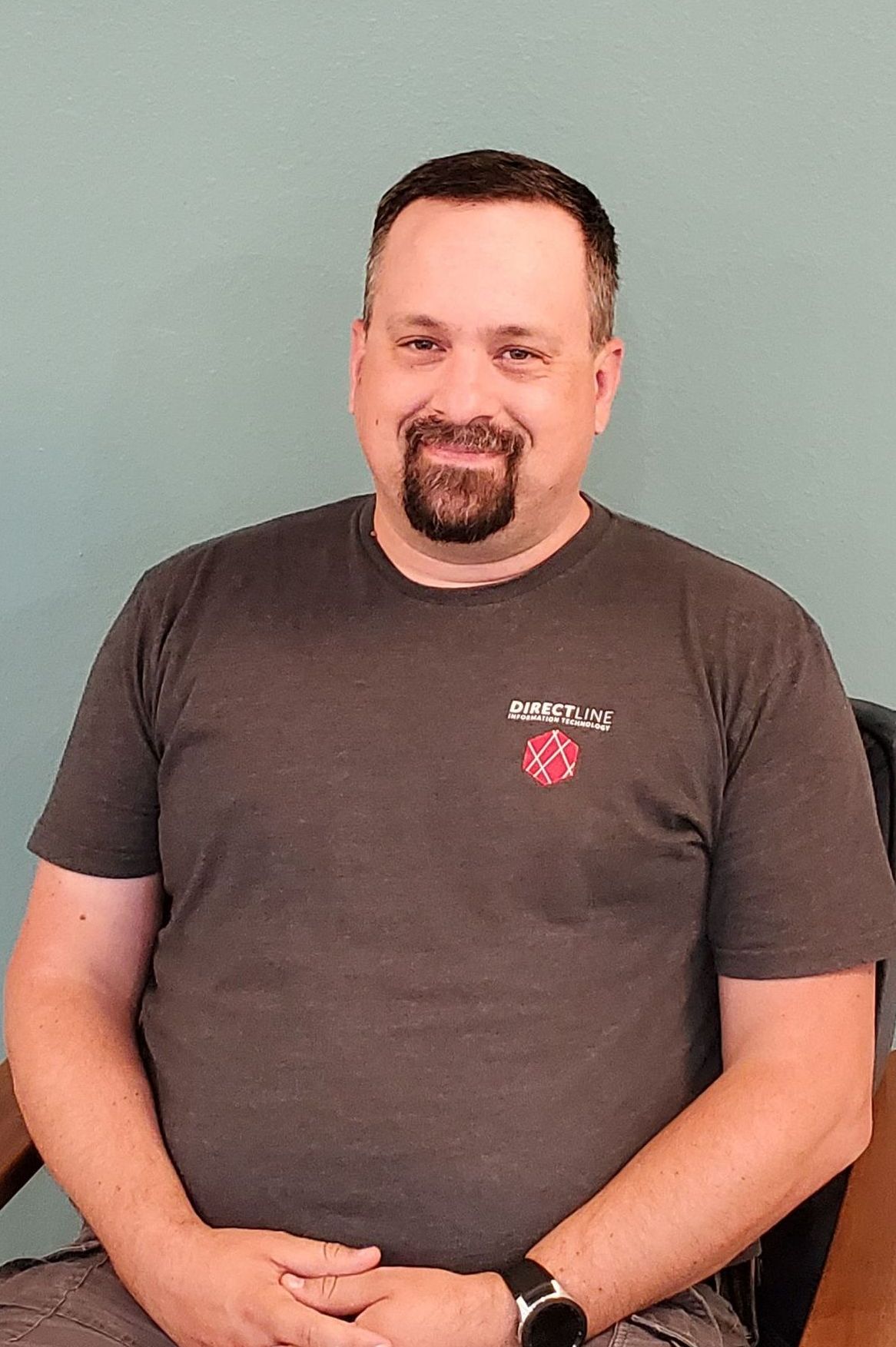 a man wearing a brown t shirt with branding of DirectLine It. Dark hair with goatee type of beard and mustache. 