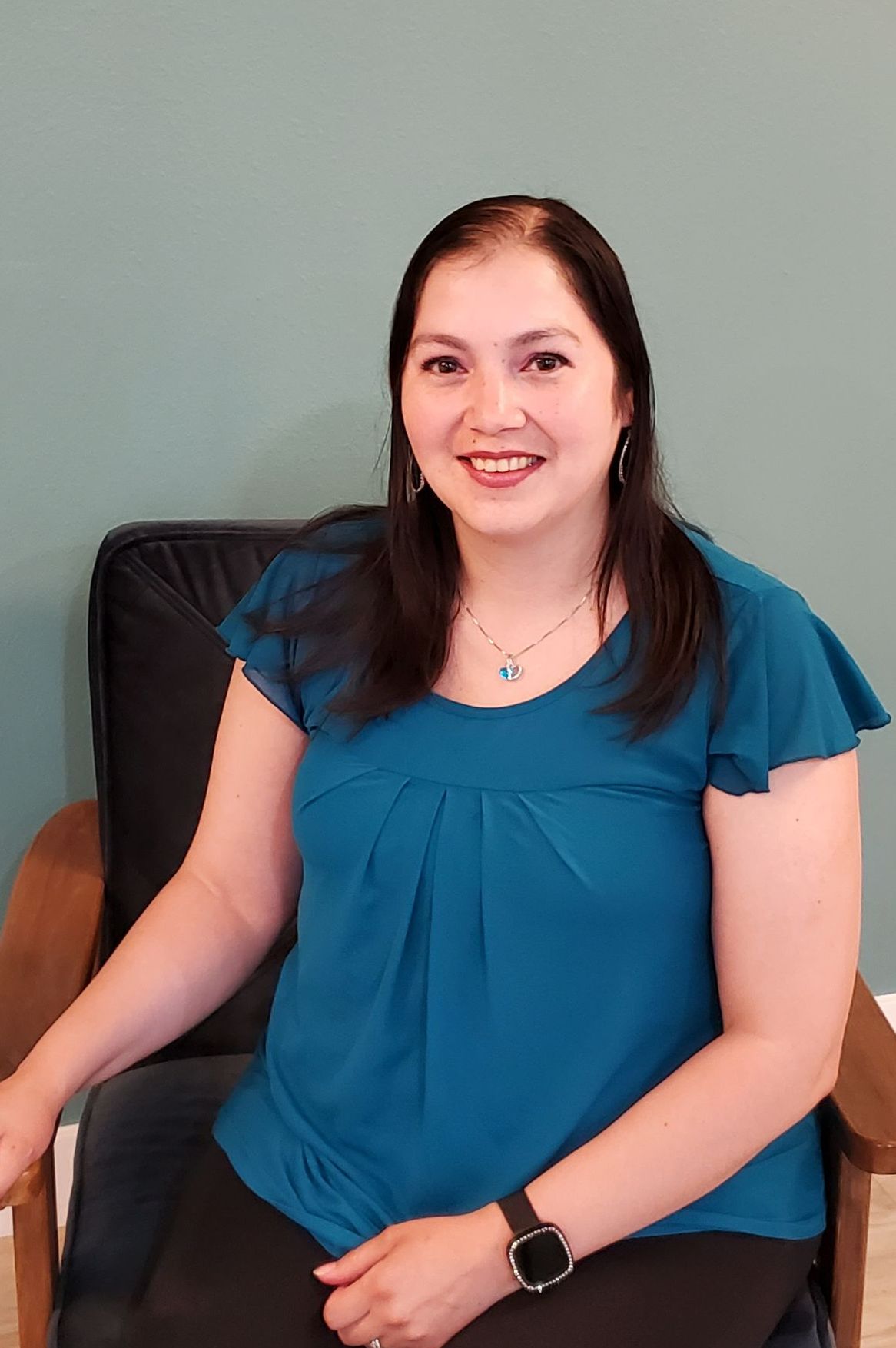 a woman sitting in a chair with dark hair and teal blouse and black slacks