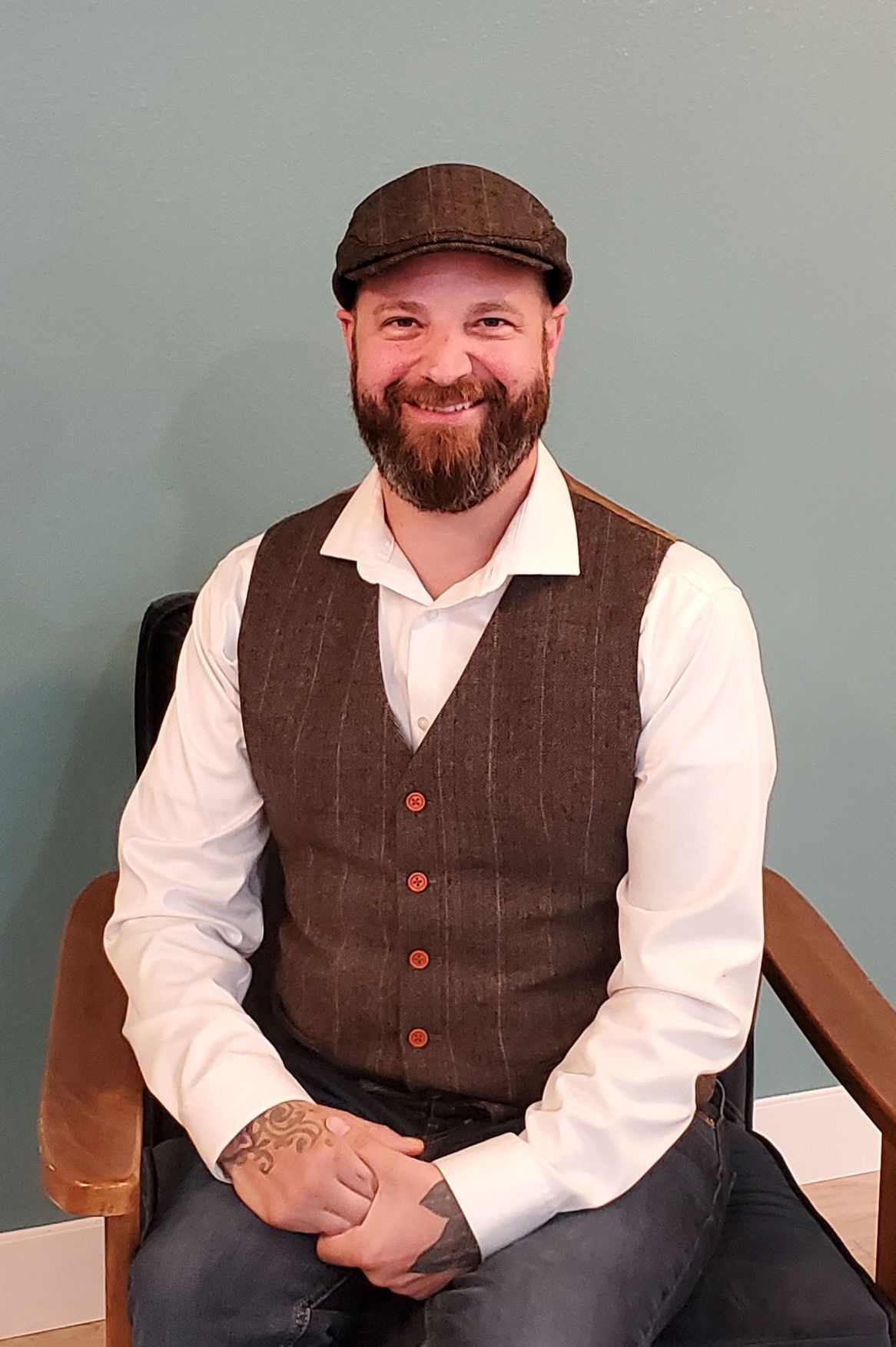 A man sitting, wearing wool vest and cap with beard and mustache smiling