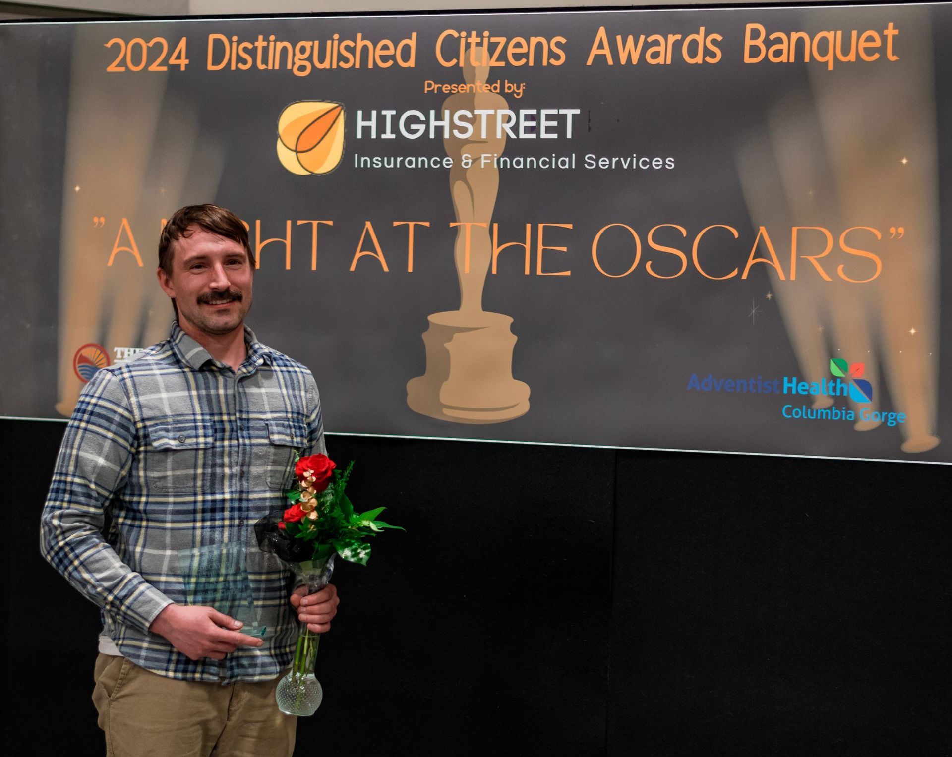 A man is holding a vase of flowers in front of a sign that says 2024 distinguished citizens awards banquet