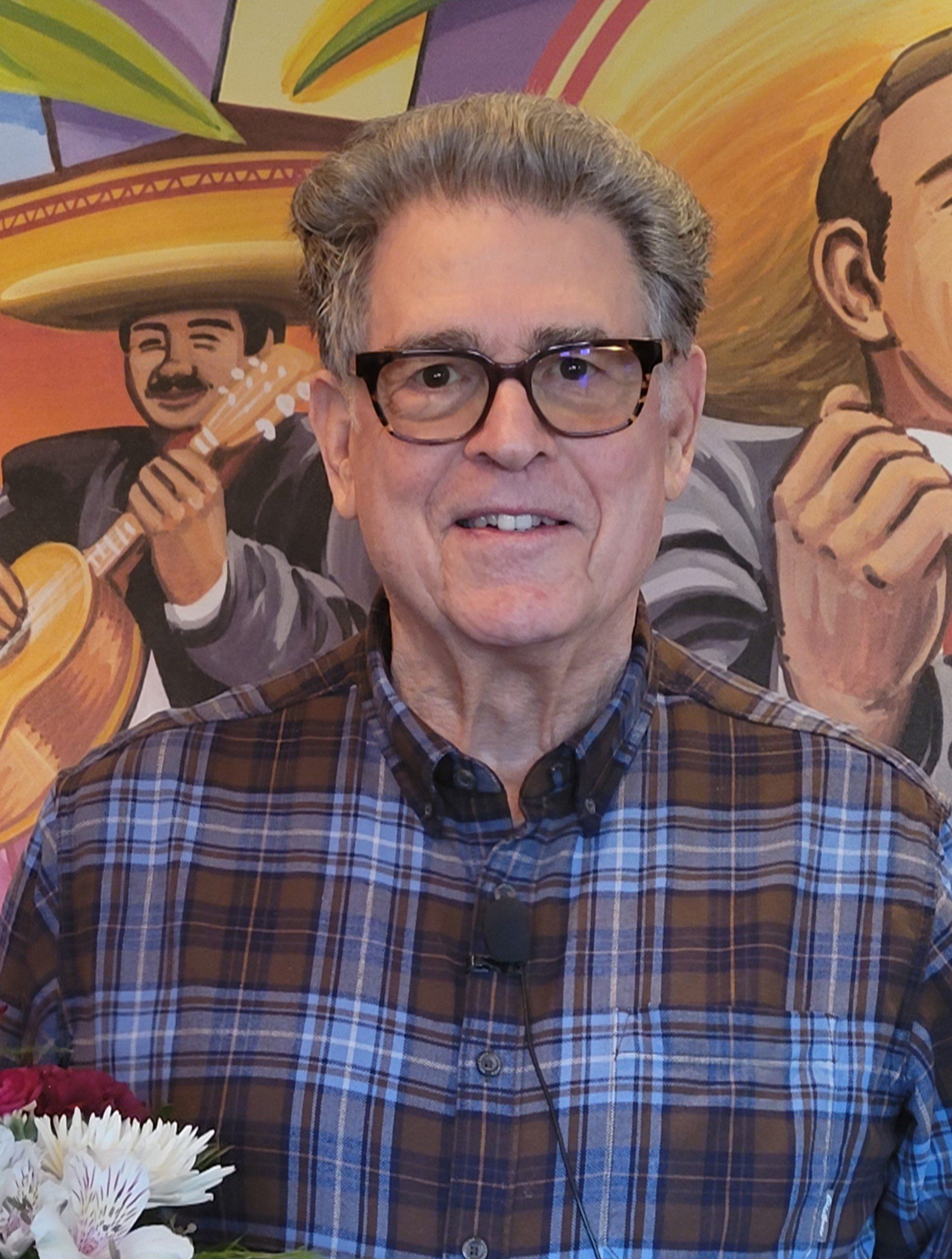 A man wearing glasses and a plaid shirt is standing in front of a painting of a man playing a guitar.