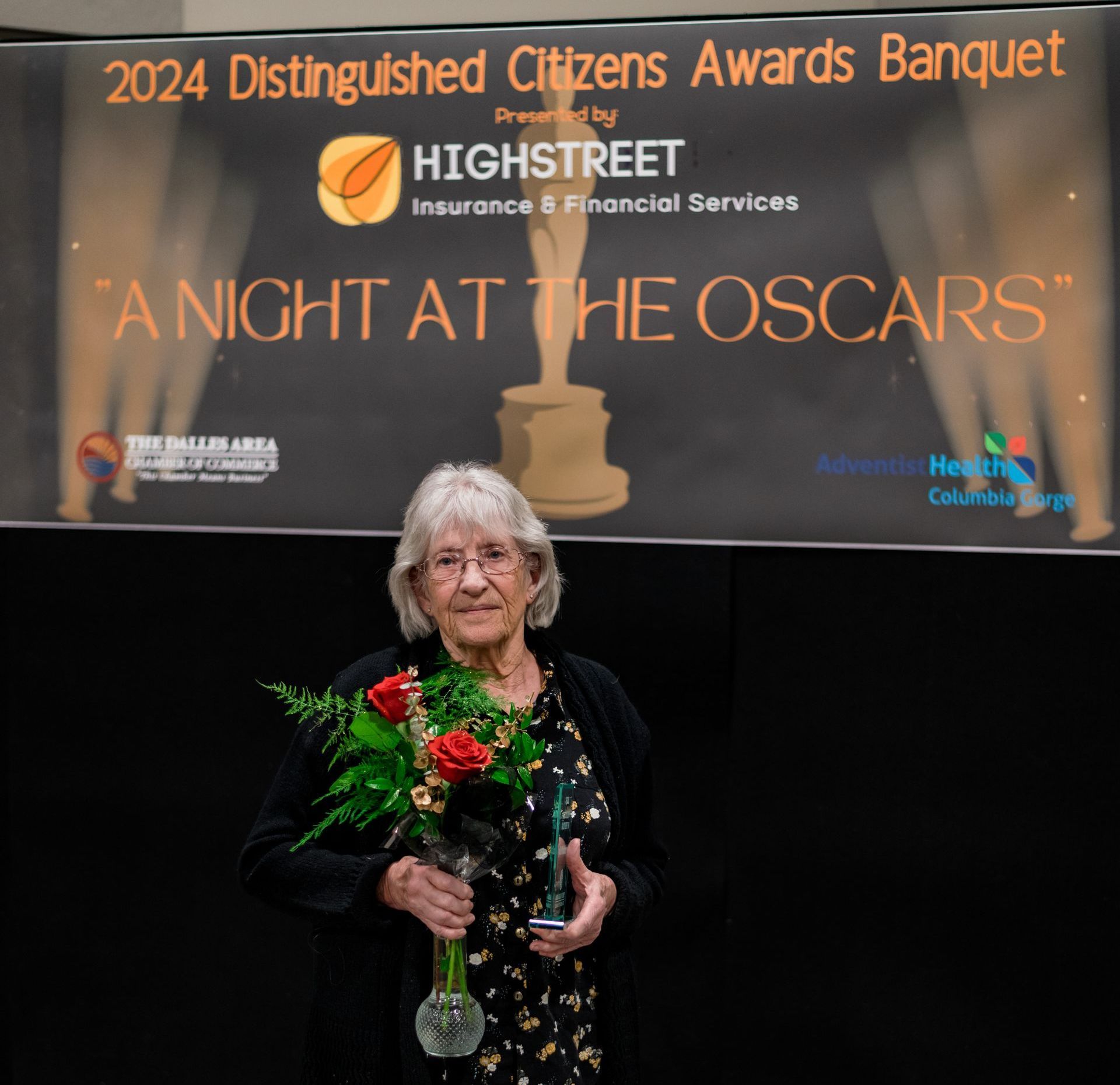 A woman is holding a vase of flowers in front of a sign that says 2024 distinguished citizens awards banquet